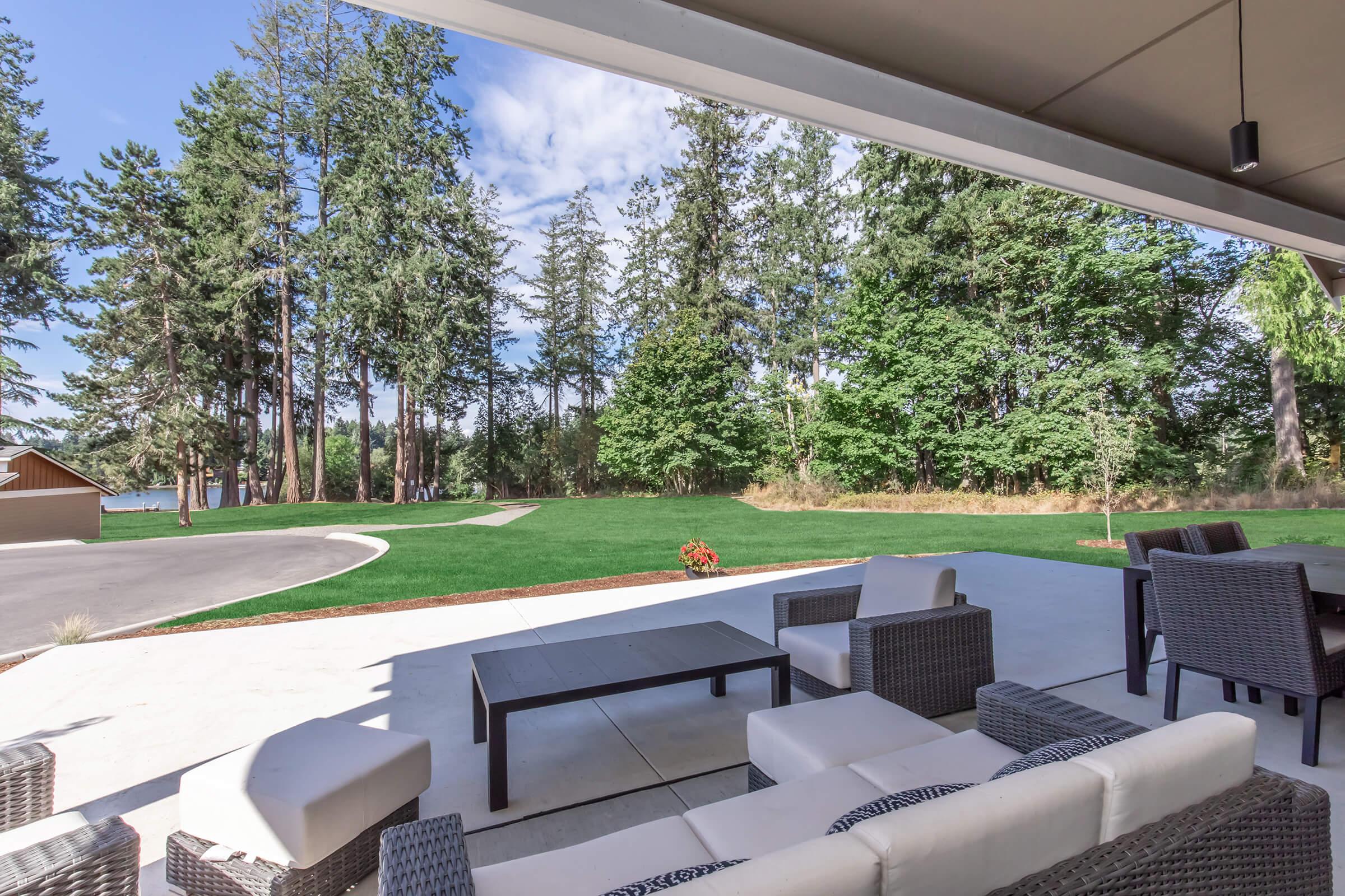 A well-furnished patio with a modern seating arrangement, featuring a white couch and a black coffee table. In the background, there is a lush green lawn bordered by tall trees under a partly cloudy sky. A winding path can be seen, creating a serene outdoor atmosphere.