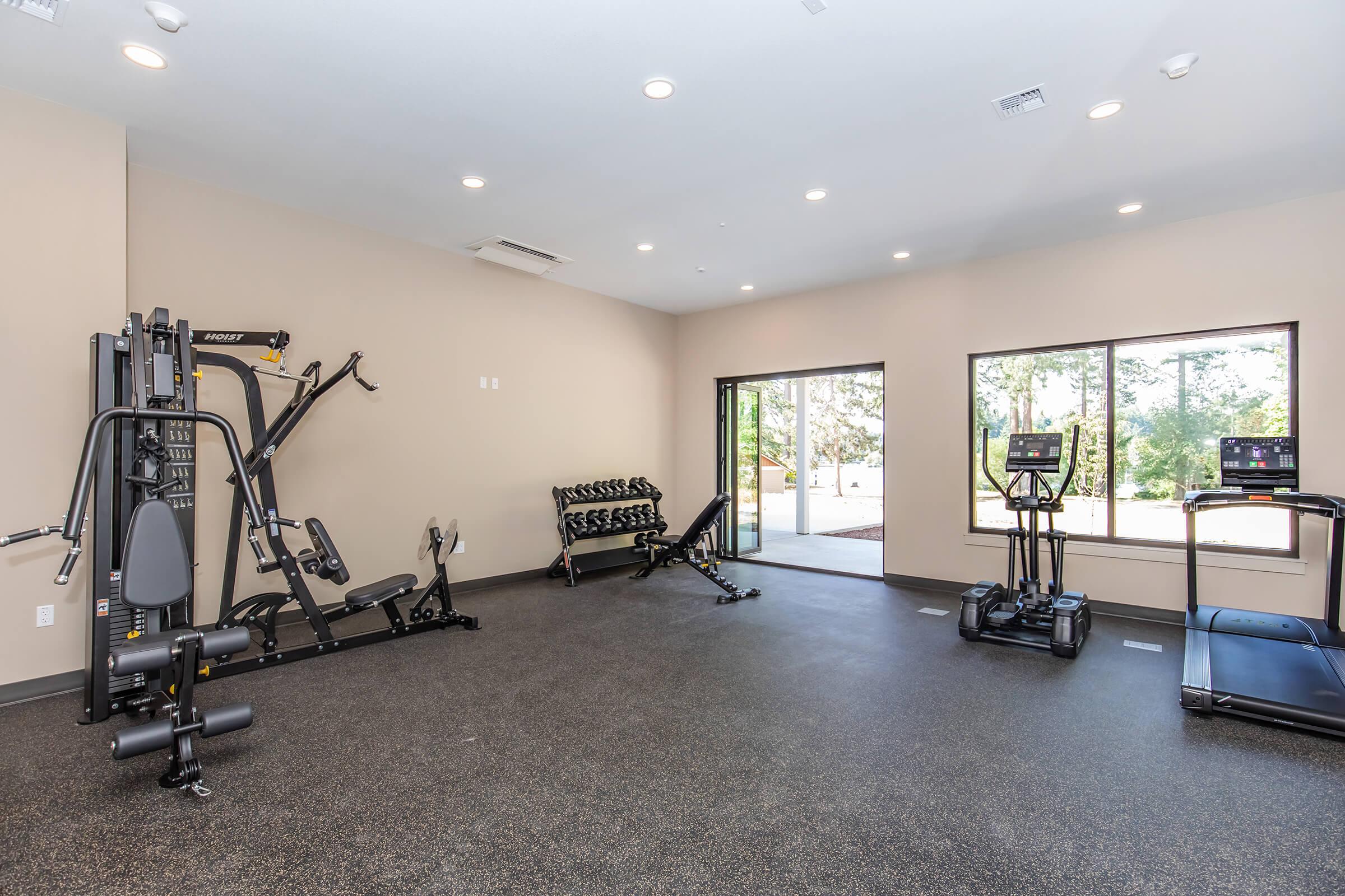 A modern gym interior featuring a multi-functional weight machine, a set of free weights on a rack, a treadmill, and an elliptical machine. Large windows allow natural light to illuminate the space, which has a clean and open design with dark flooring.