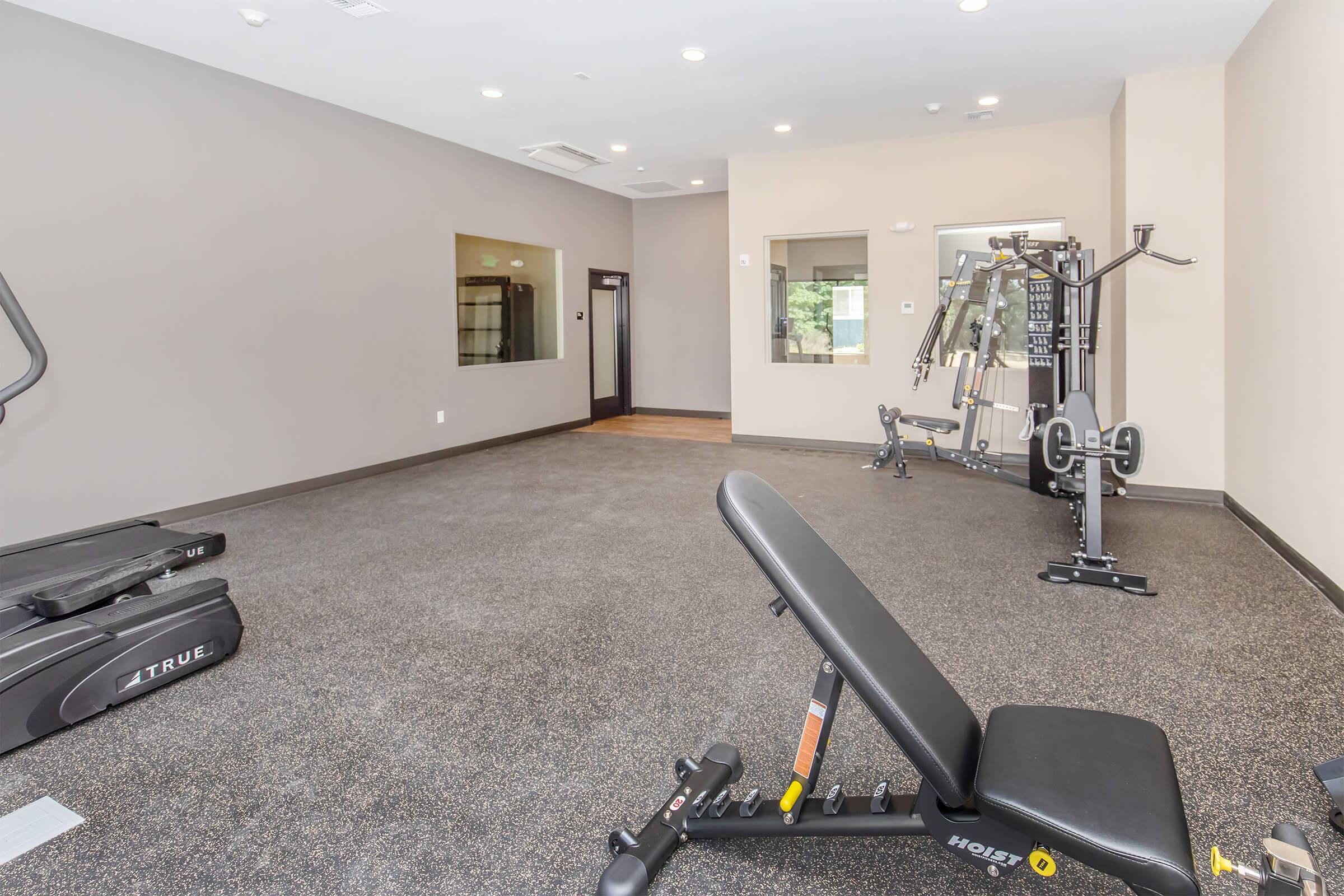 A spacious and clean gym area featuring various fitness equipment. The foreground includes an adjustable bench, while the background has a treadmill and a multi-station weight machine. The walls are painted in a neutral tone, and the flooring is black with a textured finish. Large windows allow natural light.