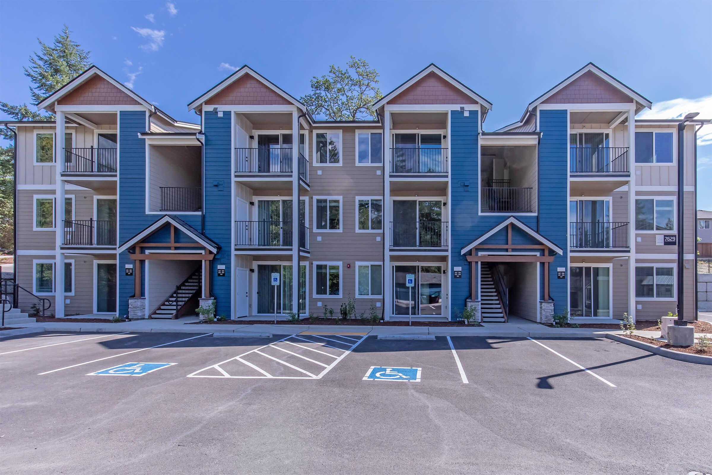 Three-story residential apartment building featuring a modern design with blue and beige siding. The building has multiple balconies, staircases, and accessible parking spaces in front. Surrounding area includes greenery and clear blue skies.