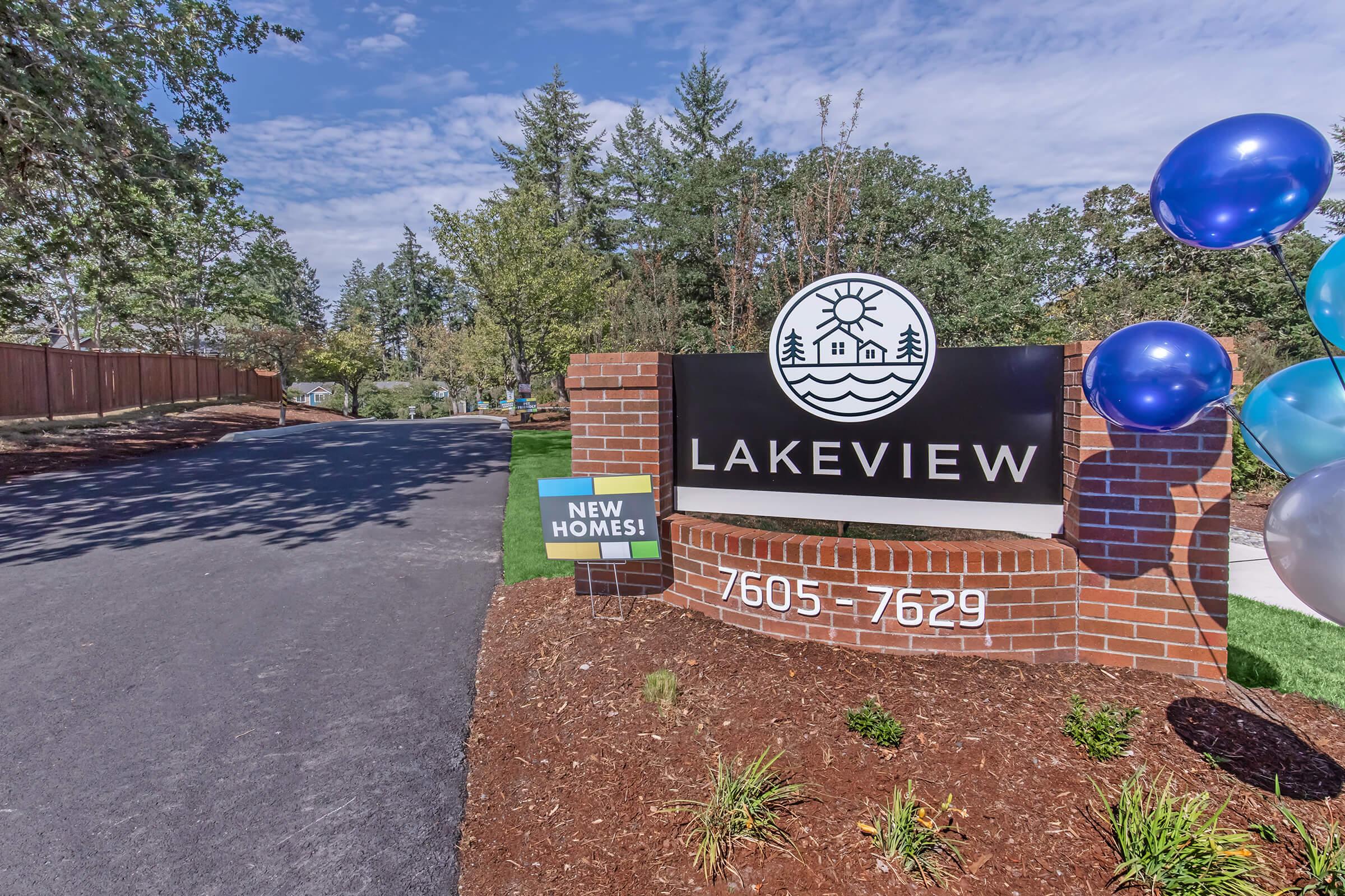 Sign for Lakeview community with a logo featuring trees and a house, accompanied by a nearby sign advertising "NEW HOMES!" and balloons in blue and white colors. The setting is landscaped, with a clear blue sky overhead.
