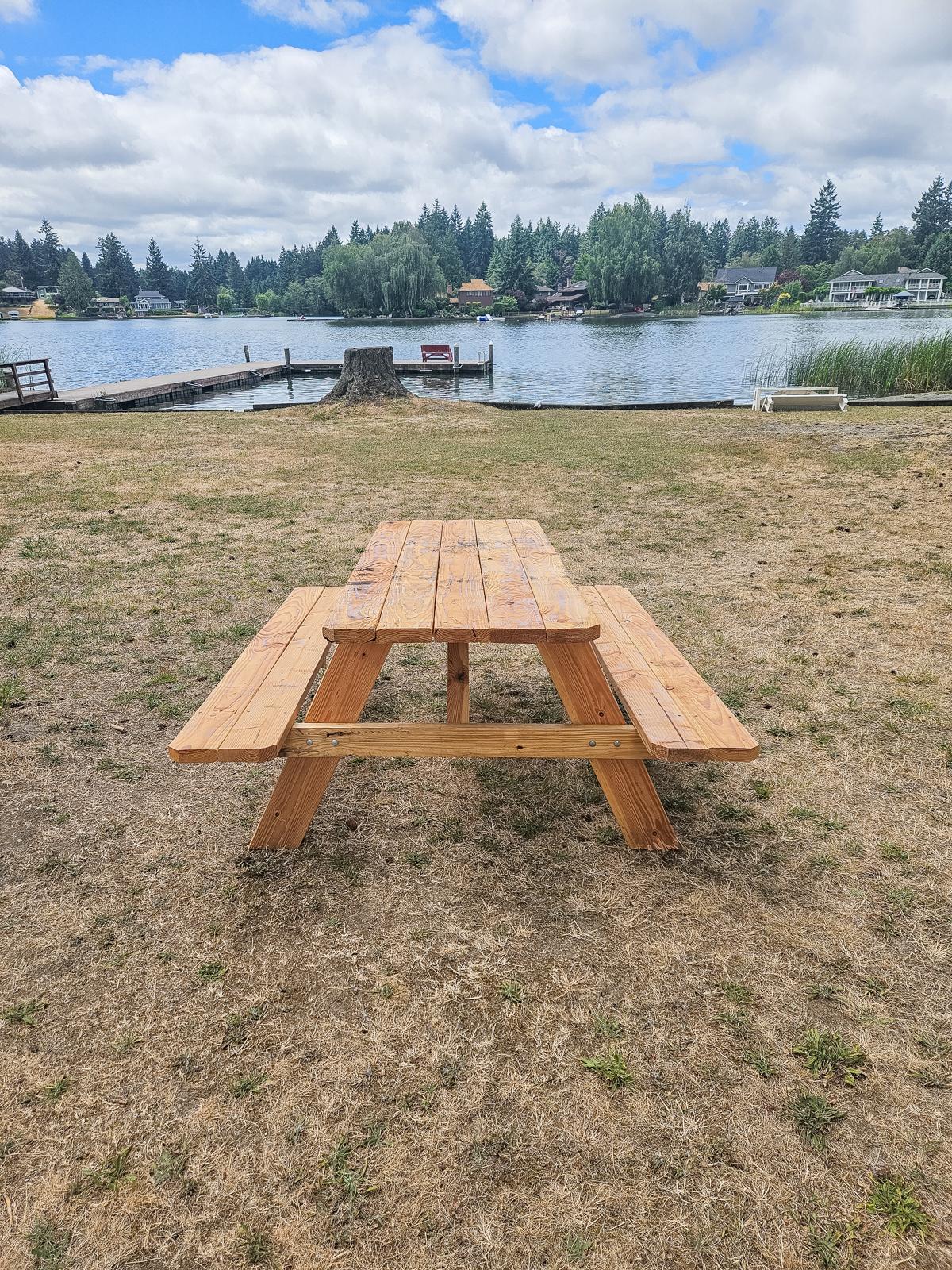 Wooden picnic table on a grassy area by a lake. The sky is partly cloudy, and there are trees and houses visible across the water in the background. The ground appears dry, indicating a warm climate.