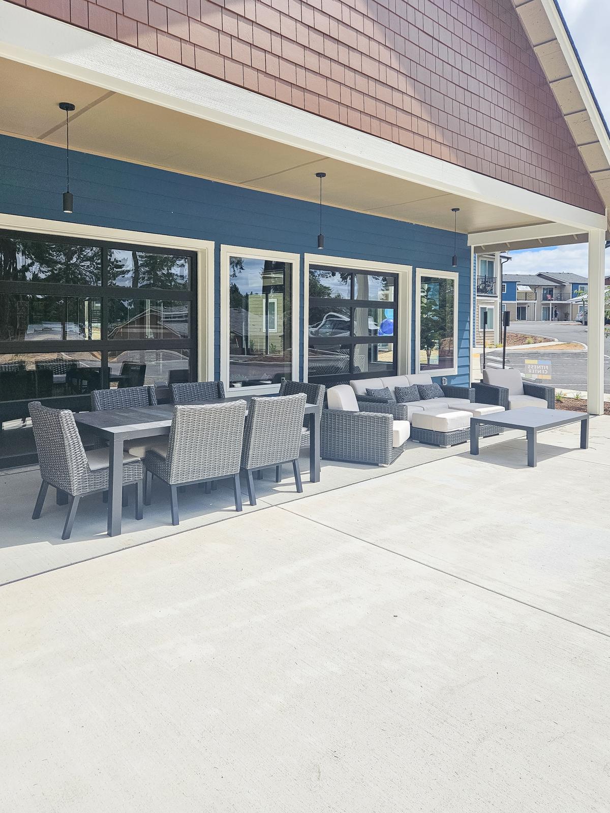Outdoor seating area featuring modern wicker furniture, including a large dining table and several lounge chairs, situated next to large windows of a building. The patio is designed for relaxation and social gatherings, with a clean concrete floor and a sunny atmosphere.