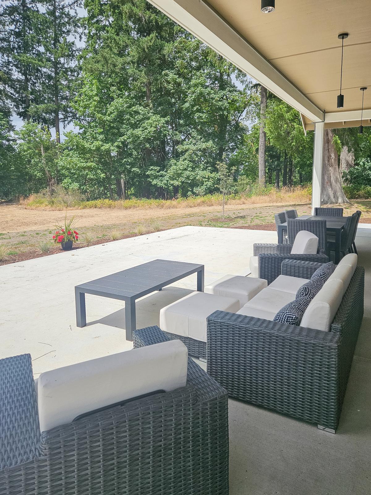 A spacious outdoor patio featuring modern wicker furniture, including a sectional sofa with white cushions, a coffee table, and a dining set. Surrounded by greenery, the area offers a serene view of trees and a grassy expanse, ideal for relaxation and social gatherings.