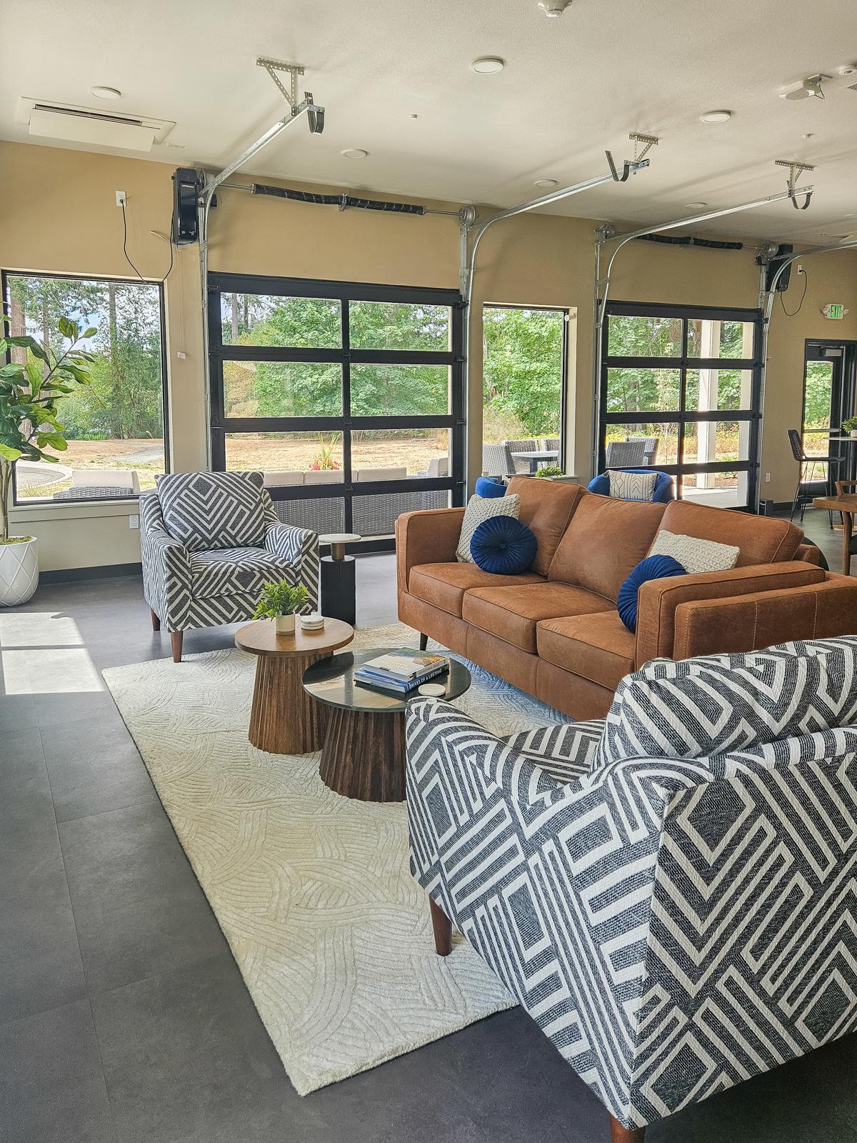 A modern lounge area featuring a brown sofa with blue accent pillows, two patterned armchairs, a round table with decor items, and a textured rug. Large windows allow natural light and offer views of greenery outside, creating a welcoming and comfortable atmosphere.