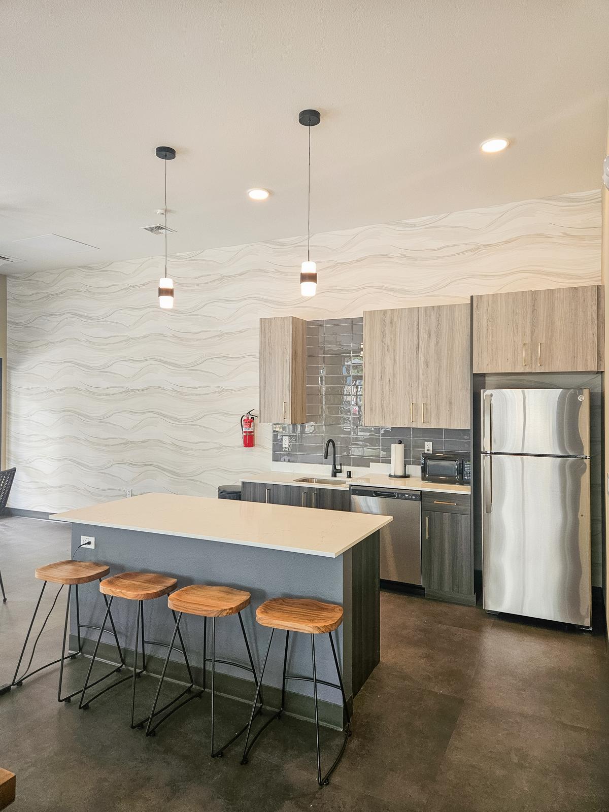 Modern kitchen interior featuring a white countertop island with four wooden bar stools. The kitchen includes stainless steel appliances, wooden cabinetry, and stylish light fixtures hanging from the ceiling. A light, wavy-patterned wall complements the sleek design.