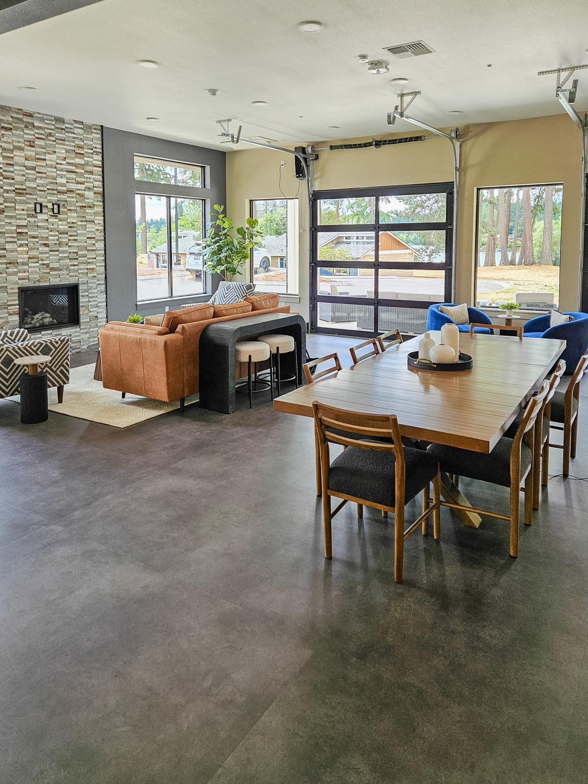 Spacious living area featuring a modern sofa, armchairs, and a decorative coffee table. A large dining table with chairs is positioned nearby. The room has large windows allowing natural light, and a stone fireplace adds a cozy touch. The floor is dark, and the decor is contemporary with a blend of textures.