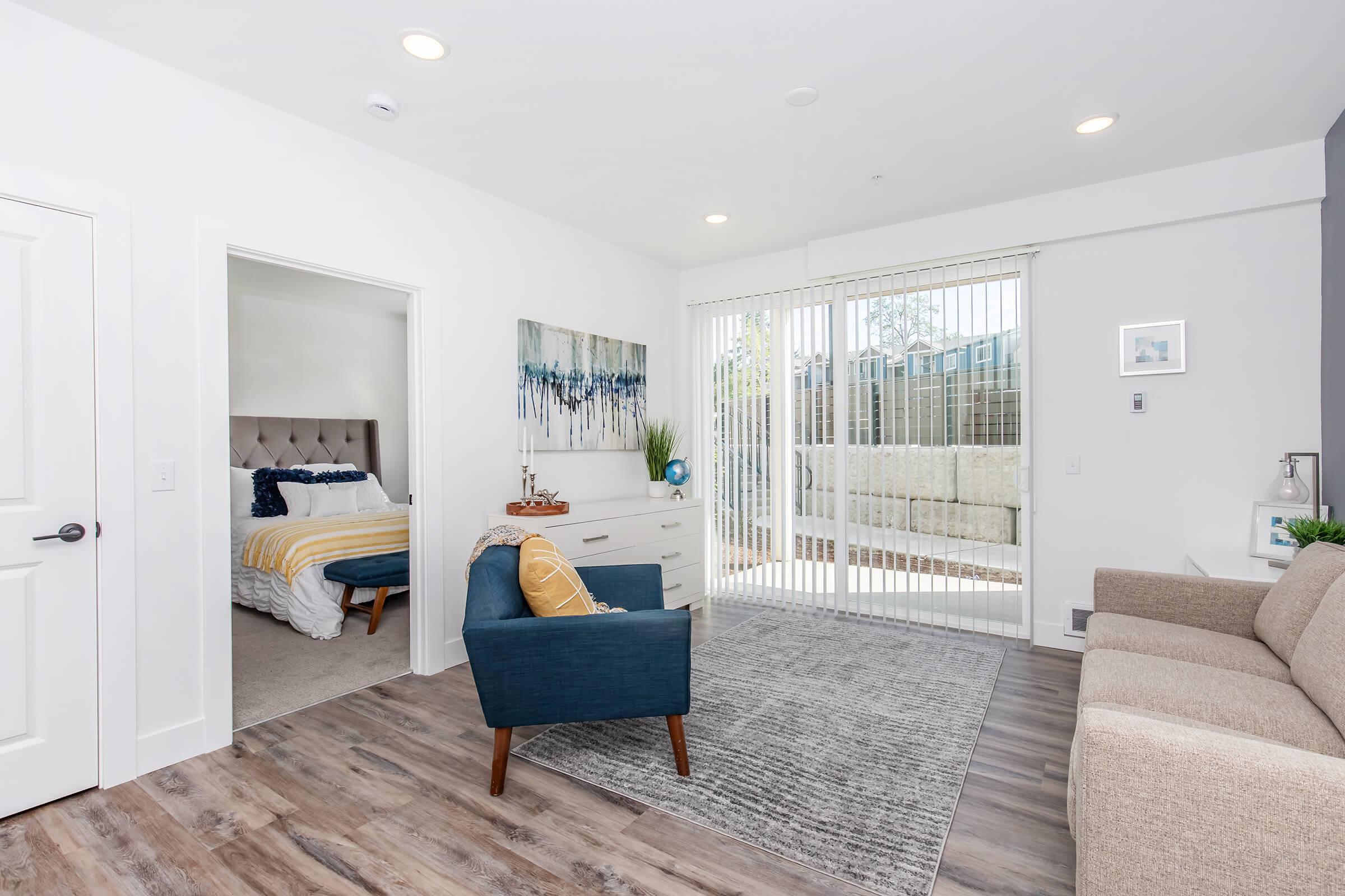 A modern, bright living room features a comfortable sofa, an accent chair, and a textured area rug. A doorway leads to a bedroom with a bed dressed in white and yellow linens. Large glass doors open to a patio area, allowing ample natural light into the space. Neutral walls and stylish decor create a welcoming atmosphere.