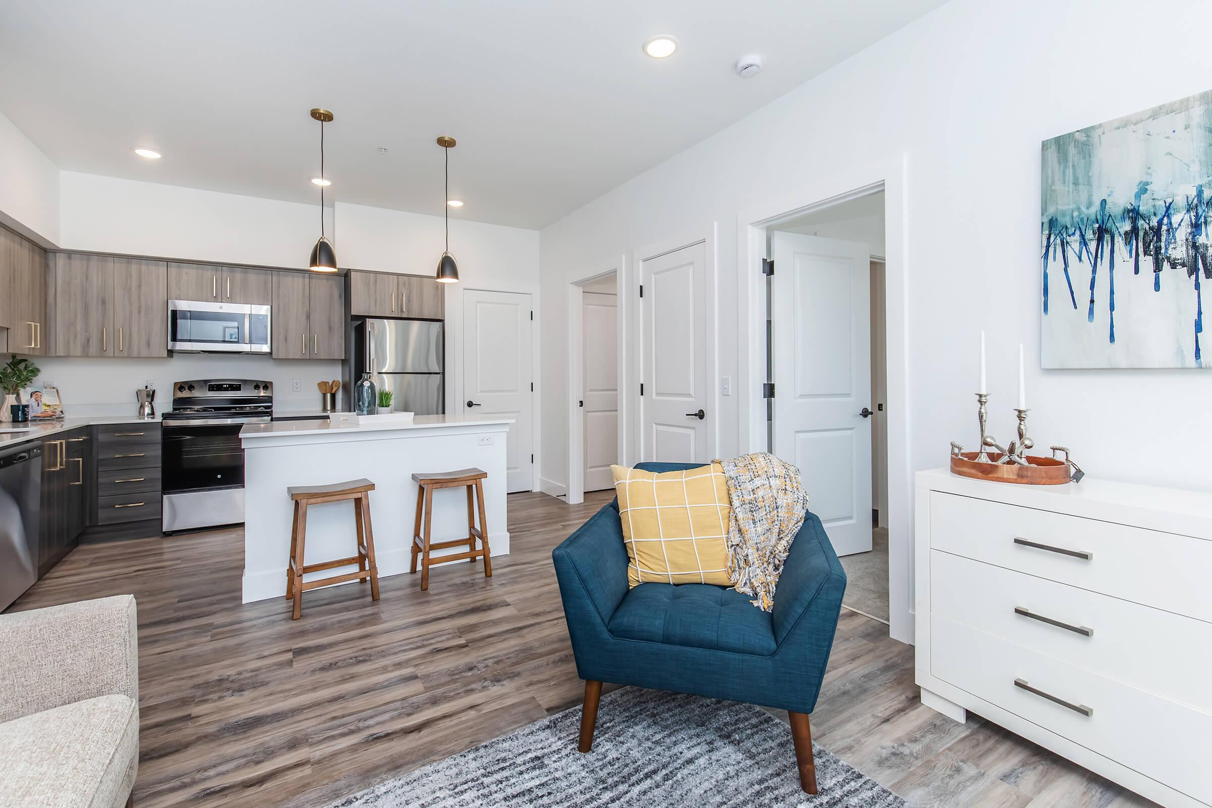 Modern open-concept living area featuring a kitchen with stainless steel appliances, a bar with two stools, and a cozy seating area with a blue chair and a yellow throw blanket. Light wood cabinets and a white decorative piece on the wall add a contemporary touch, while large windows provide natural light.
