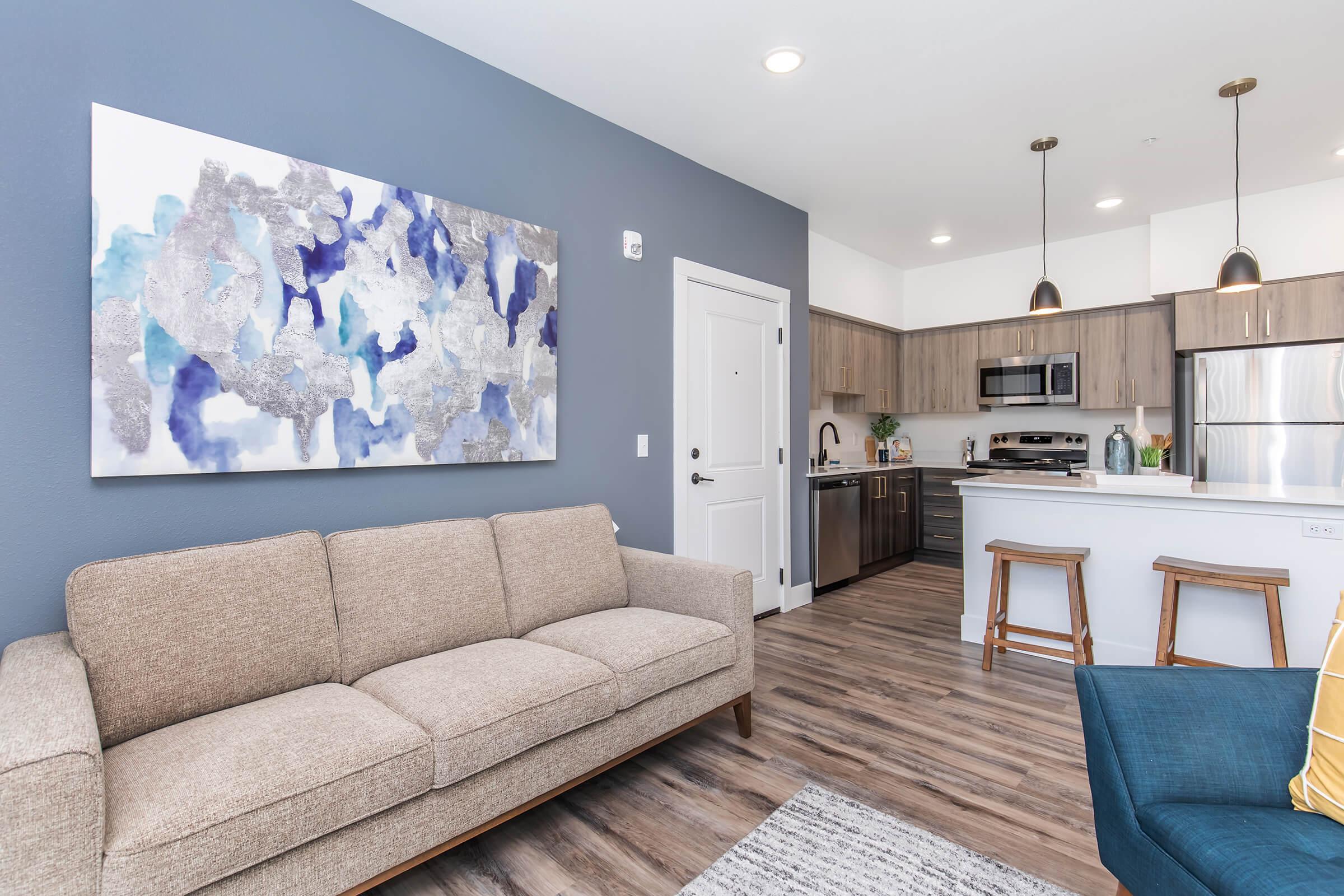 A modern living room featuring a beige sofa, a blue accent chair, and a large piece of abstract wall art with blue and grey tones. The open kitchen area in the background has wooden cabinets, stainless steel appliances, and a white countertop. Soft lighting enhances the inviting atmosphere.
