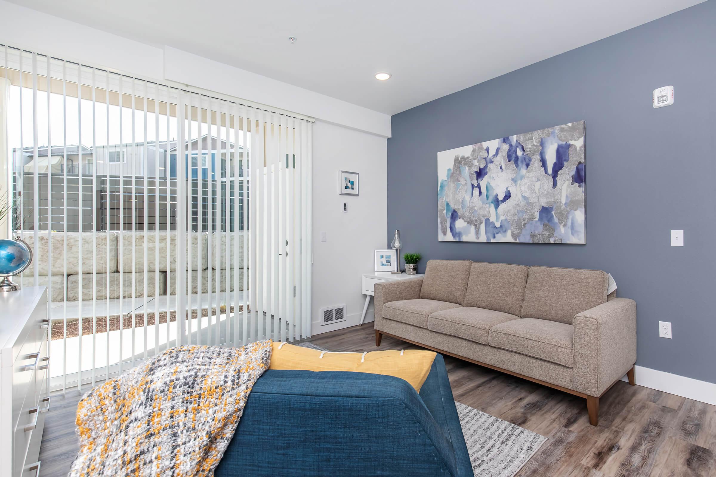 A modern living room featuring a light brown sofa, a blue accent chair with a throw blanket, a white side table, and a colorful abstract wall art. Large sliding glass doors with vertical blinds provide natural light, and a small decorative globe sits on the table. The walls are painted in soft gray and white tones.