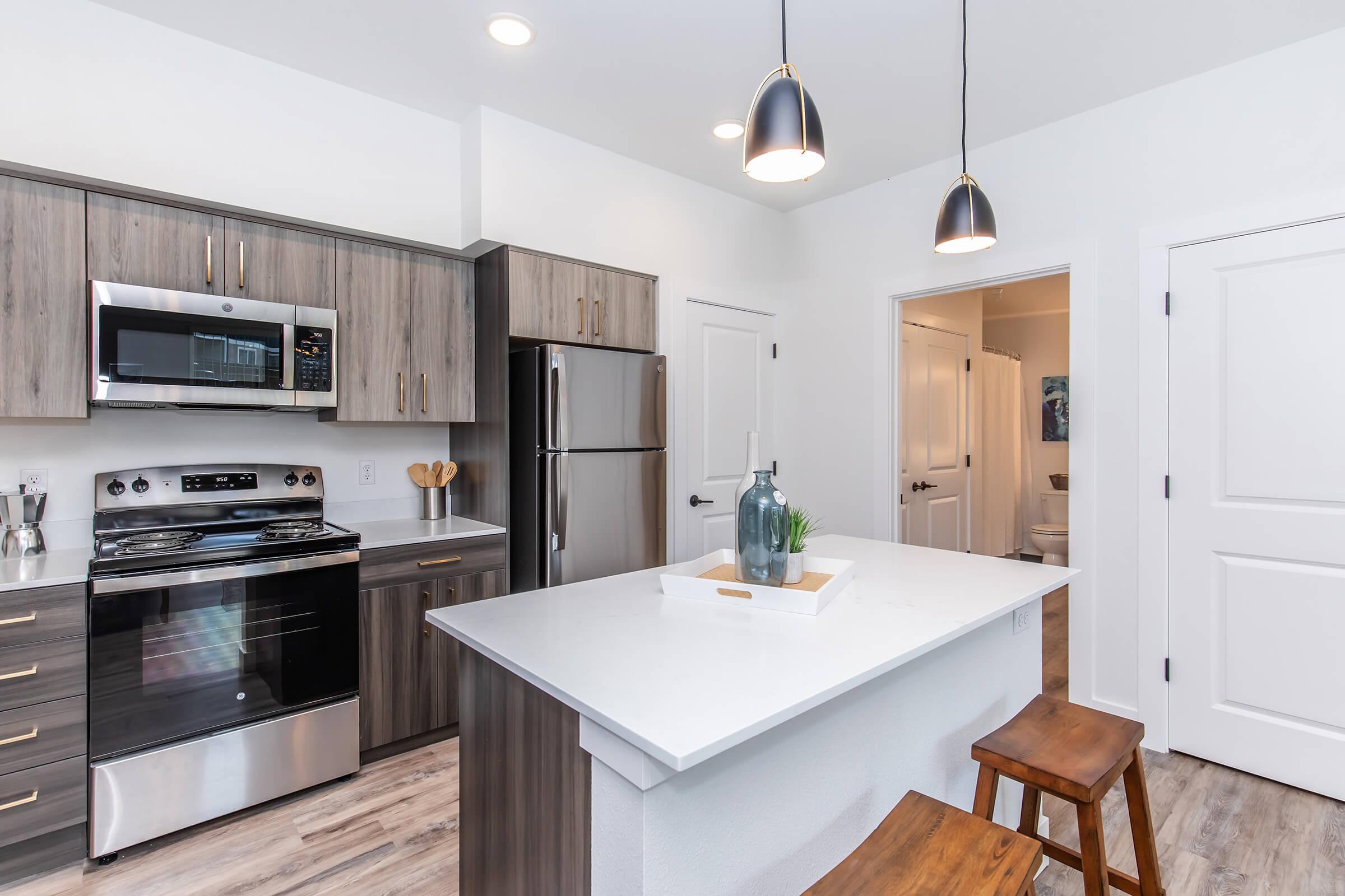 Modern kitchen featuring stainless steel appliances, including a microwave and oven, dark wood cabinets, and a white countertop. An island with two wooden bar stools is in the foreground. Soft lighting from pendant lights illuminates the space, and there is a door leading to an adjacent room.