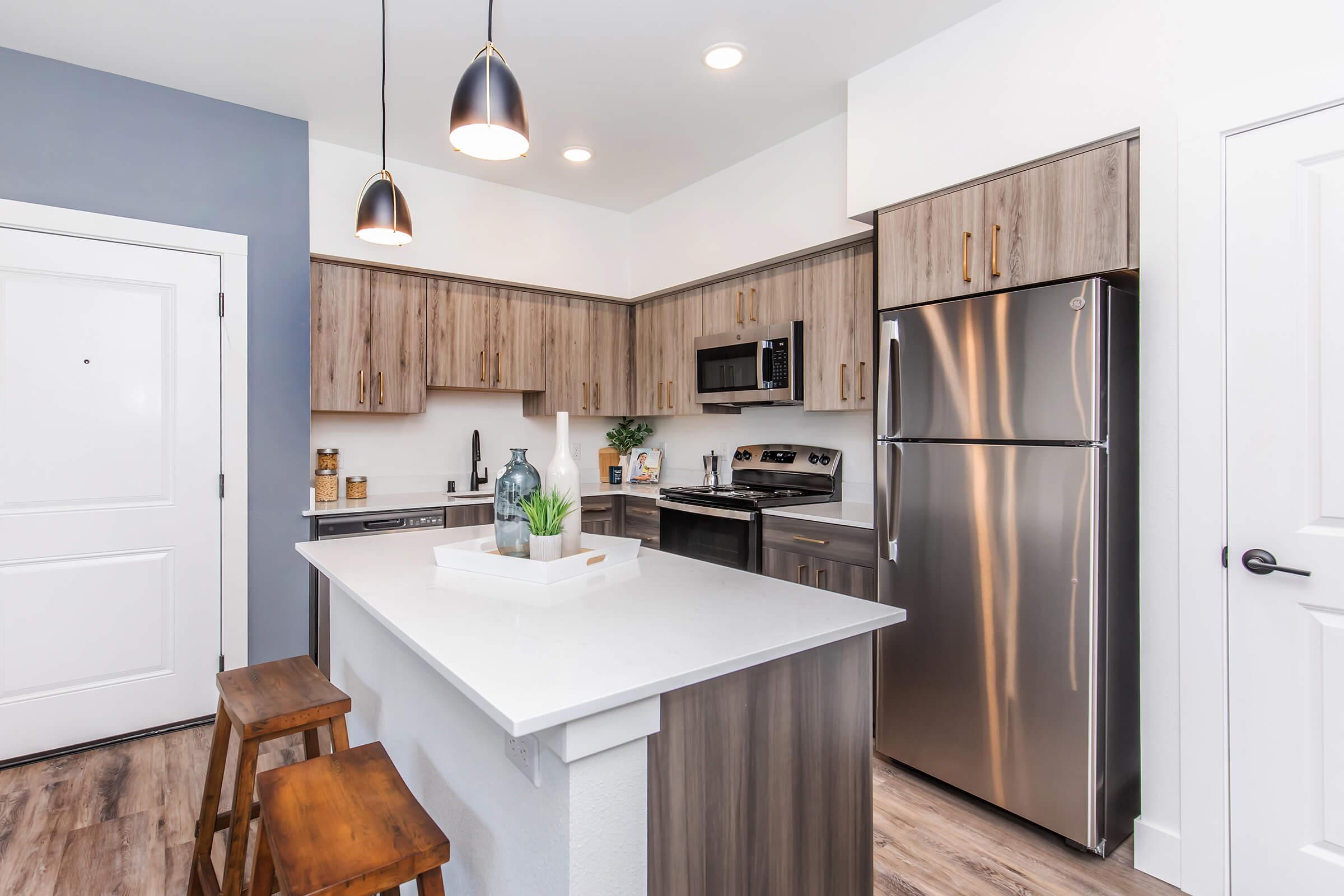 Modern kitchen featuring wooden cabinets, stainless steel appliances, and a central island with seating. The space includes pendant lighting, a microwave, and a refrigerator, with a plant on the island for decor. The walls are painted in a light gray tone, creating a bright and inviting atmosphere.