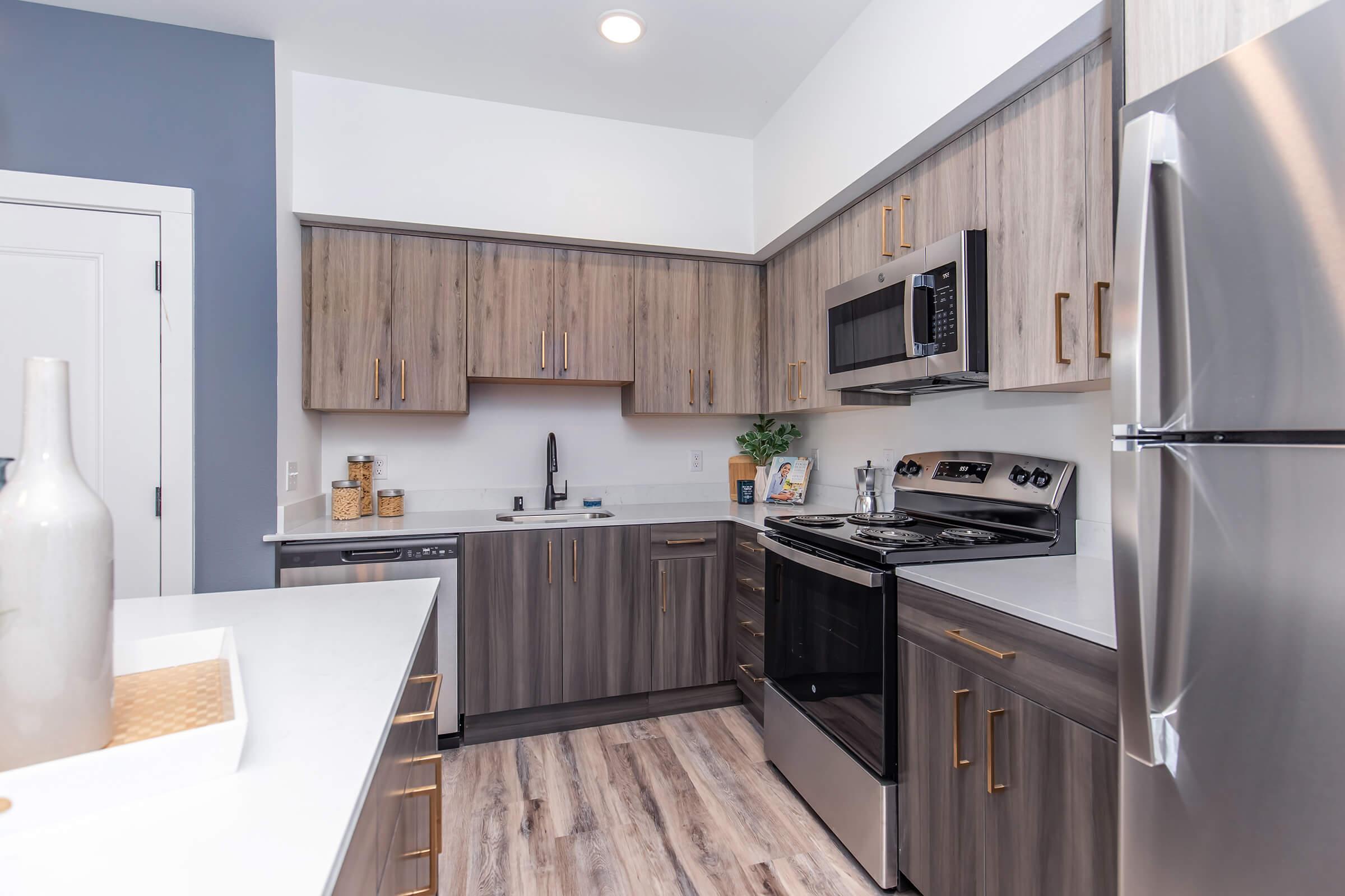 Modern kitchen featuring wooden cabinets in a mix of light and dark tones. The countertop is white with a sink and stylish faucet. Stainless steel appliances include a microwave and stove. A decorative vase sits on the counter, and there are canisters and a small plant for added decor. The walls are painted in soft colors.