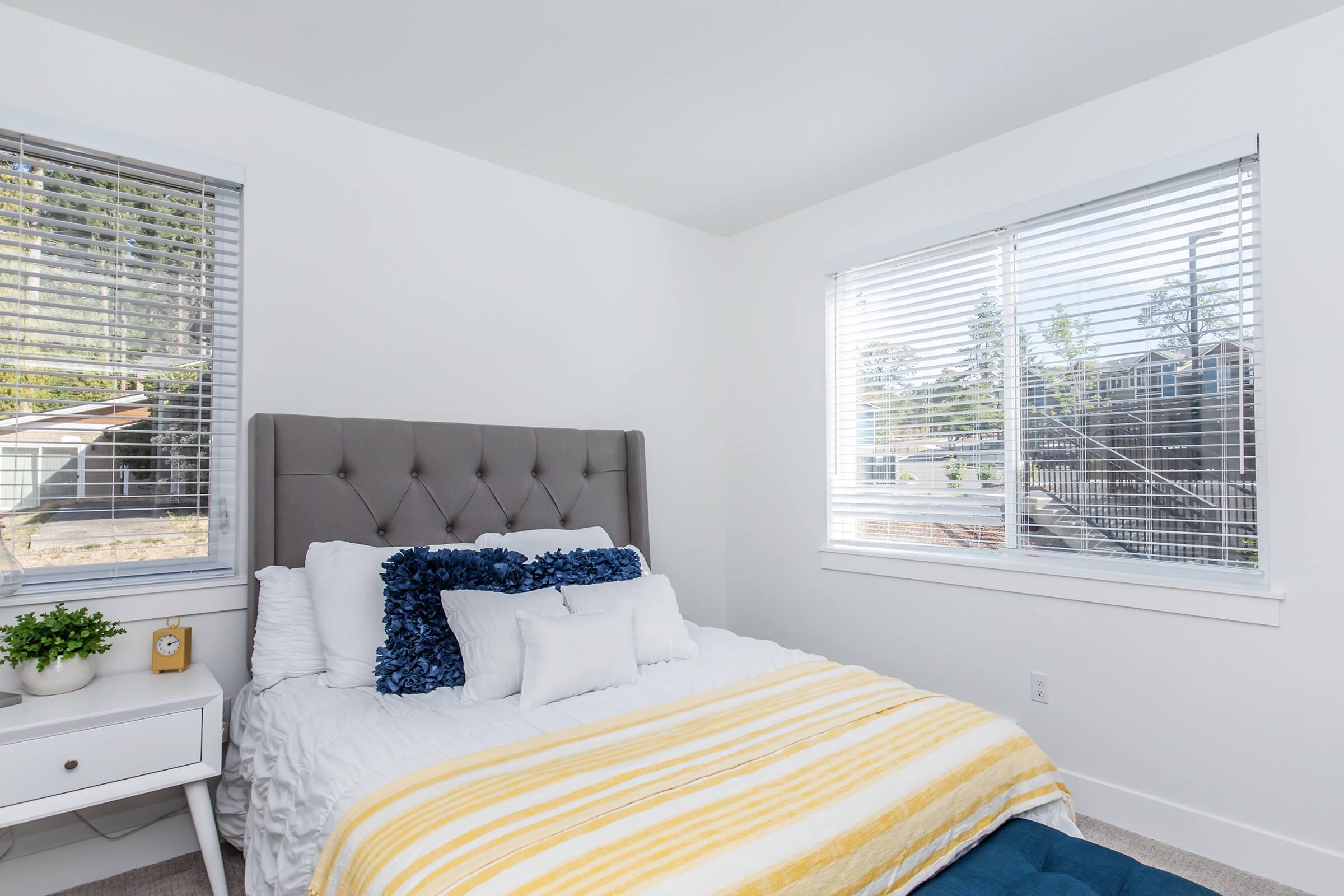 A well-lit bedroom featuring a gray upholstered bed with white and yellow bedding, decorative pillows, and a small nightstand with a plant. Two windows with blinds let in natural light, revealing a serene outdoor view. The walls are painted white, creating a clean and modern atmosphere.
