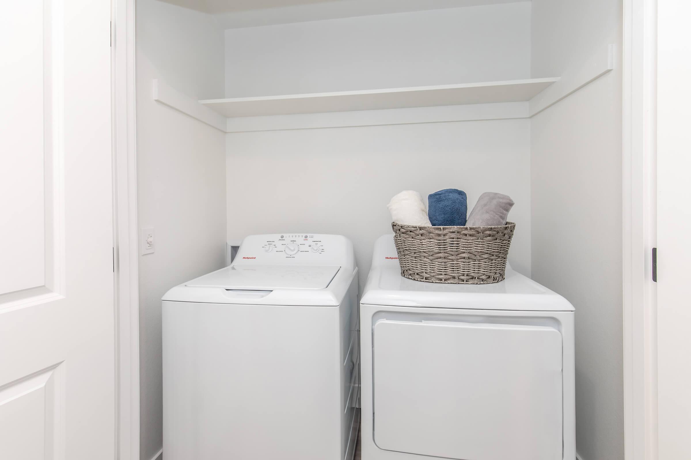 A clean laundry room featuring a white washing machine and dryer side by side. Above, there’s a shelf with no items, and next to the machines, a woven basket holds neatly folded towels in various colors. The walls and doors are painted white, creating a bright and tidy space.