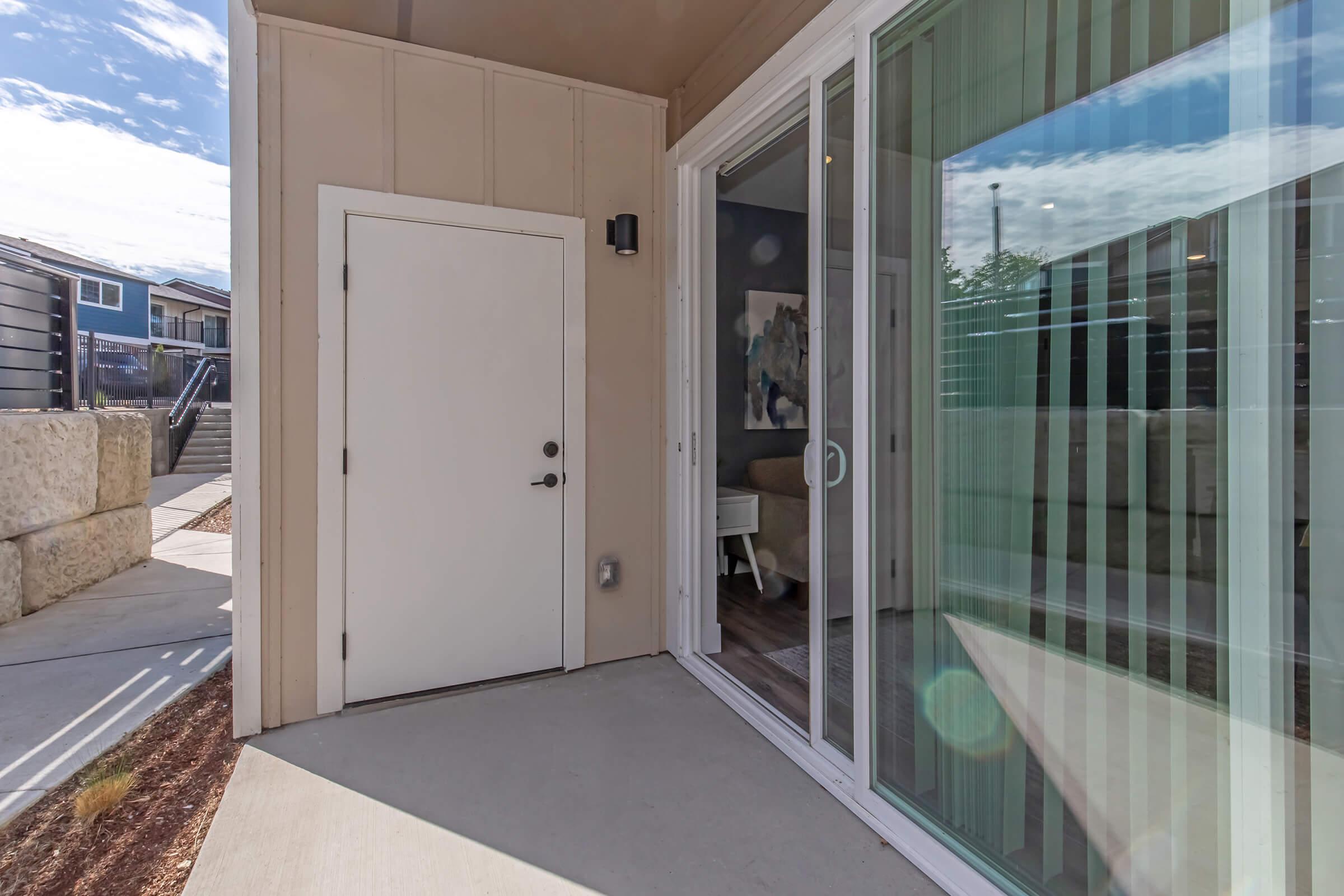 An entranceway featuring a white door and large glass sliding doors. Outside, a patio area is visible, bordered by light-colored walls. Natural light illuminates the space, highlighting a modern and inviting atmosphere.