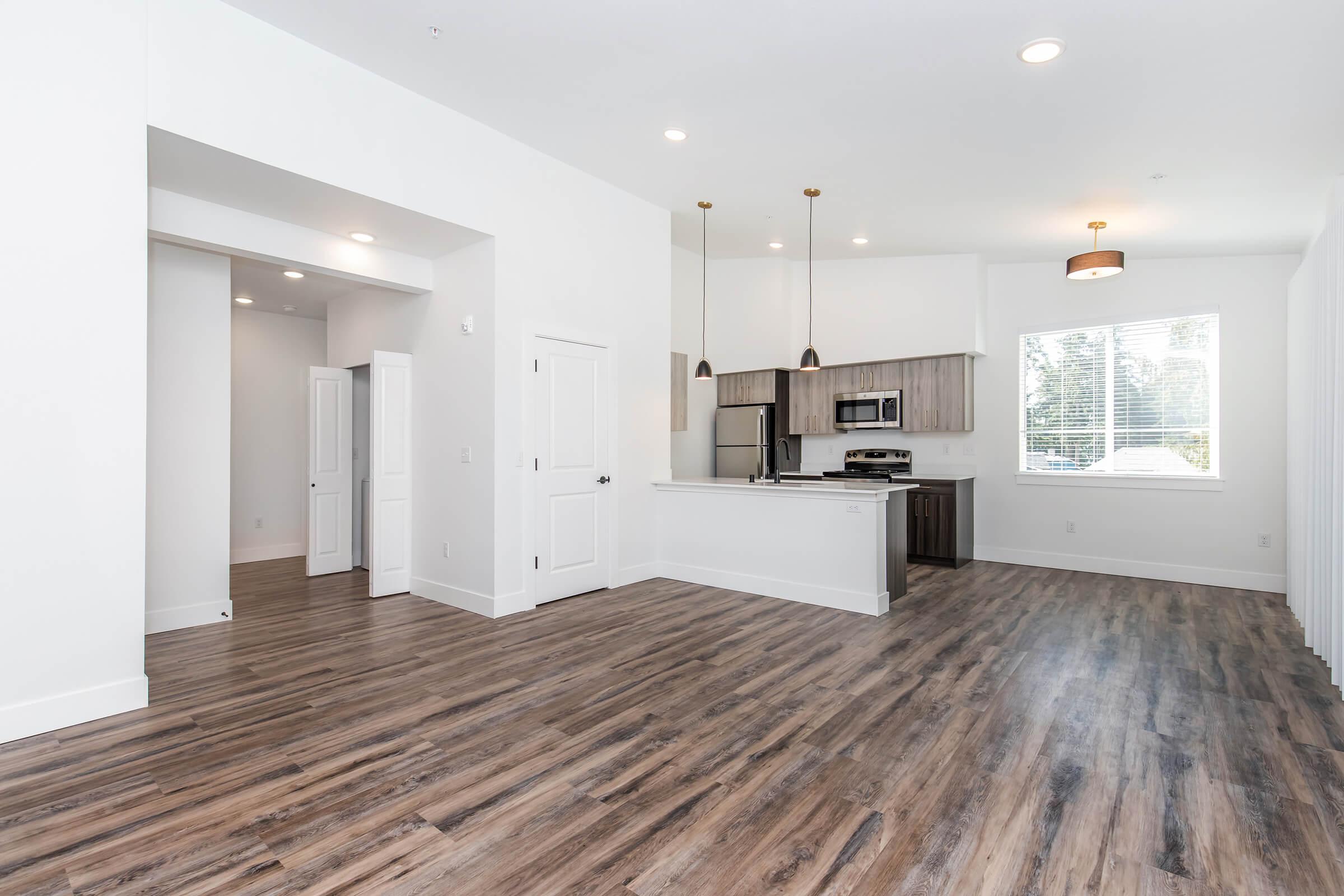 A spacious, modern living area featuring light-colored walls and wood-style flooring. The open layout includes a kitchen with stainless steel appliances and pendant lighting. Large windows allow natural light, and a wide doorway leads to another room, creating a bright and inviting atmosphere.