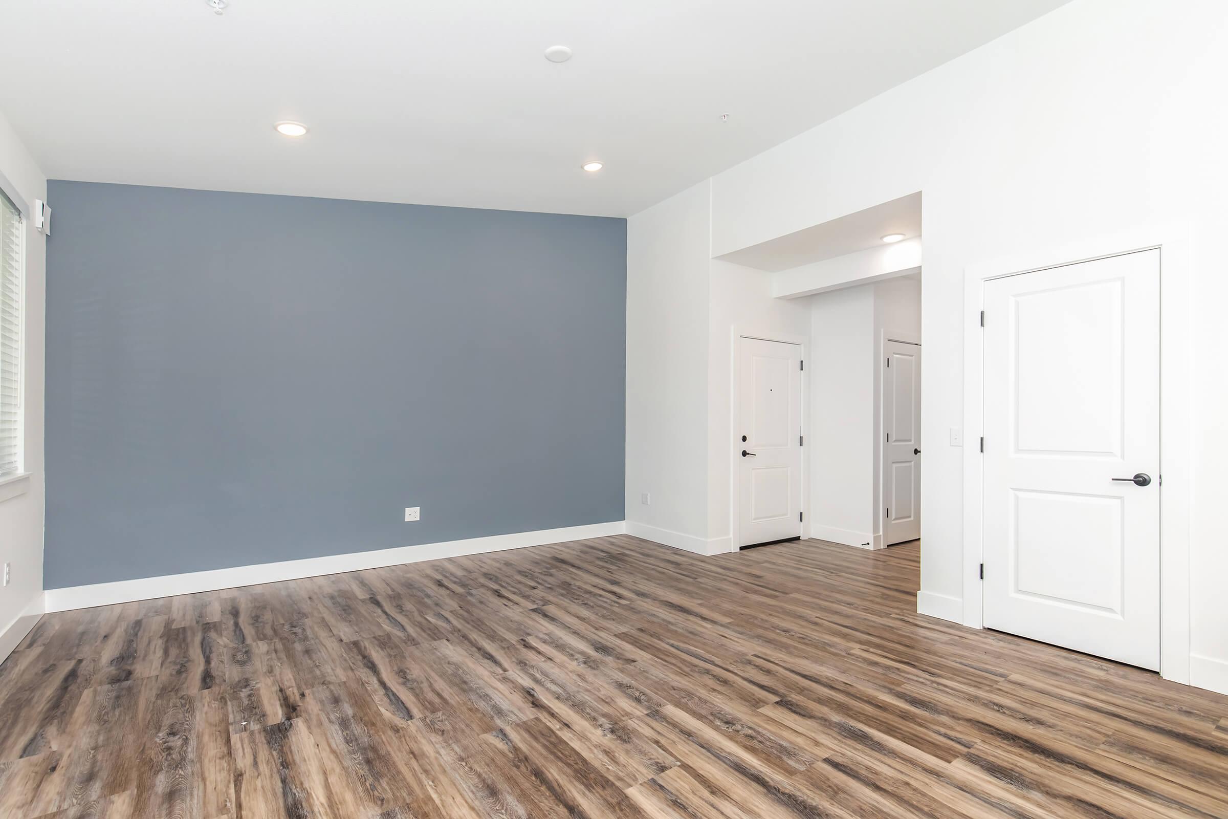 A spacious, empty room featuring wooden flooring, a light gray accent wall, and white walls. Two doors are visible on the right, leading to different areas, and ample natural light fills the space. The ceiling has modern lighting fixtures.
