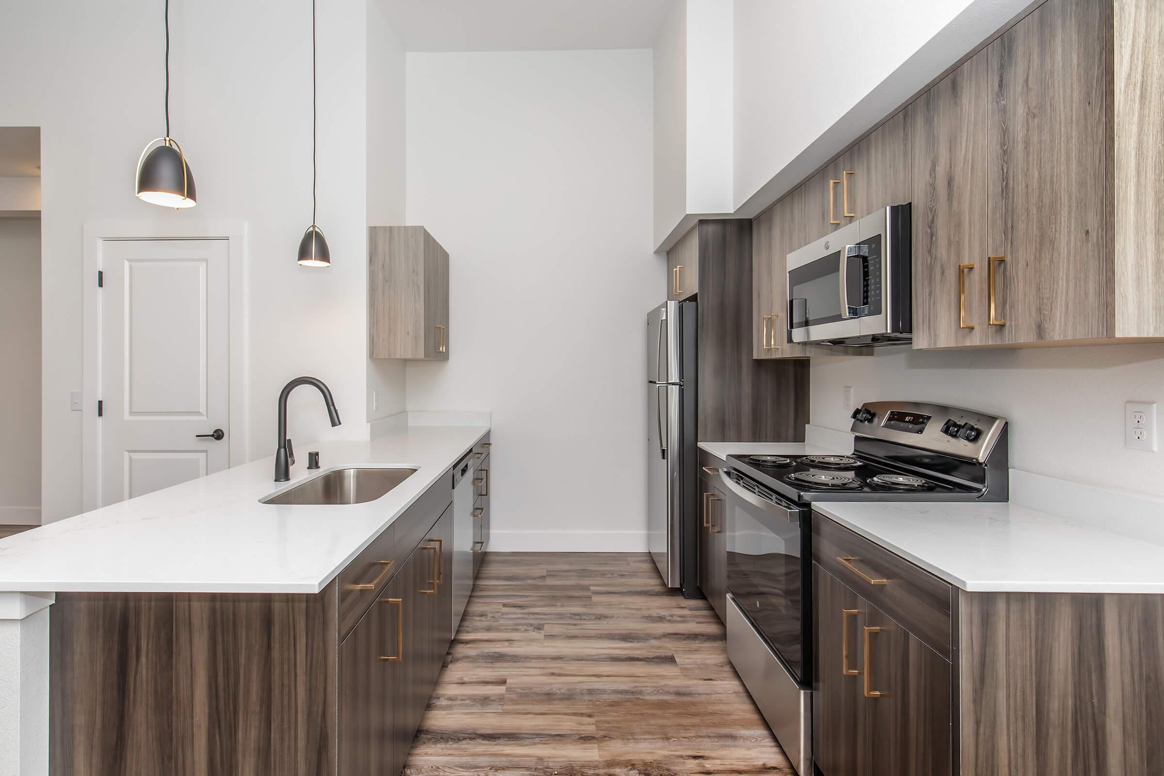 A modern kitchen featuring sleek gray cabinets with gold handles, stainless steel appliances including a microwave and oven, a single basin sink, and white countertops. The space is illuminated by stylish pendant lights, and has a light wood floor, creating a contemporary and inviting atmosphere.