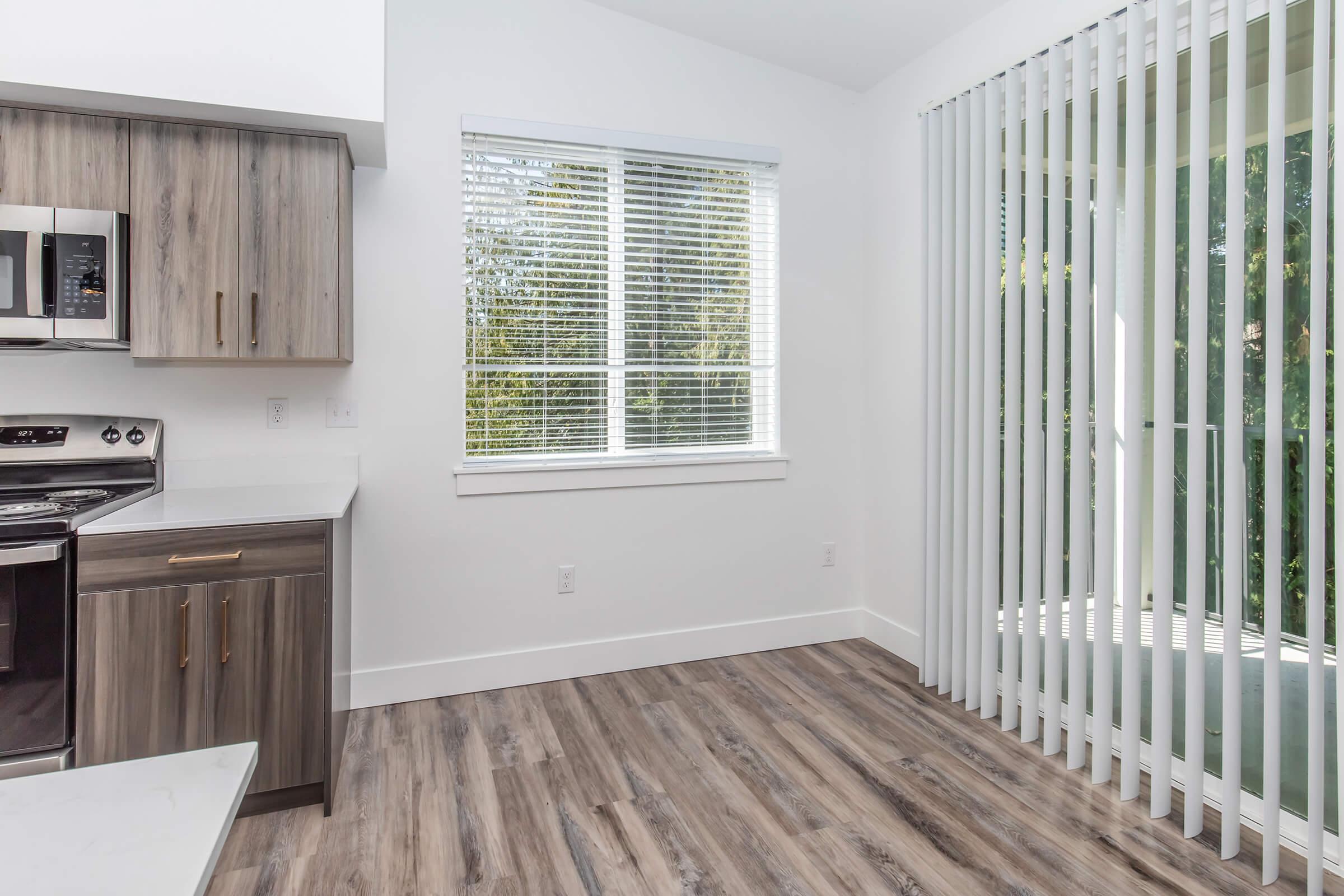 A modern kitchen space featuring stainless steel appliances, wooden cabinets, and a white countertop. Sunlight streams through large windows covered with vertical blinds, and the floor is finished with laminate wood. The area is bright and inviting, suitable for dining or relaxation.