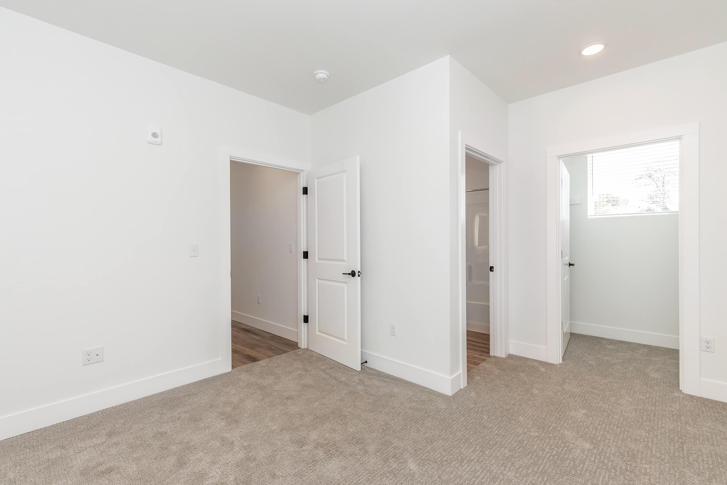 A bright, spacious interior of a room featuring light-colored walls, a soft carpeted floor, and multiple doorways leading to adjacent rooms. Natural light streams in through a window, enhancing the open feel of the space.