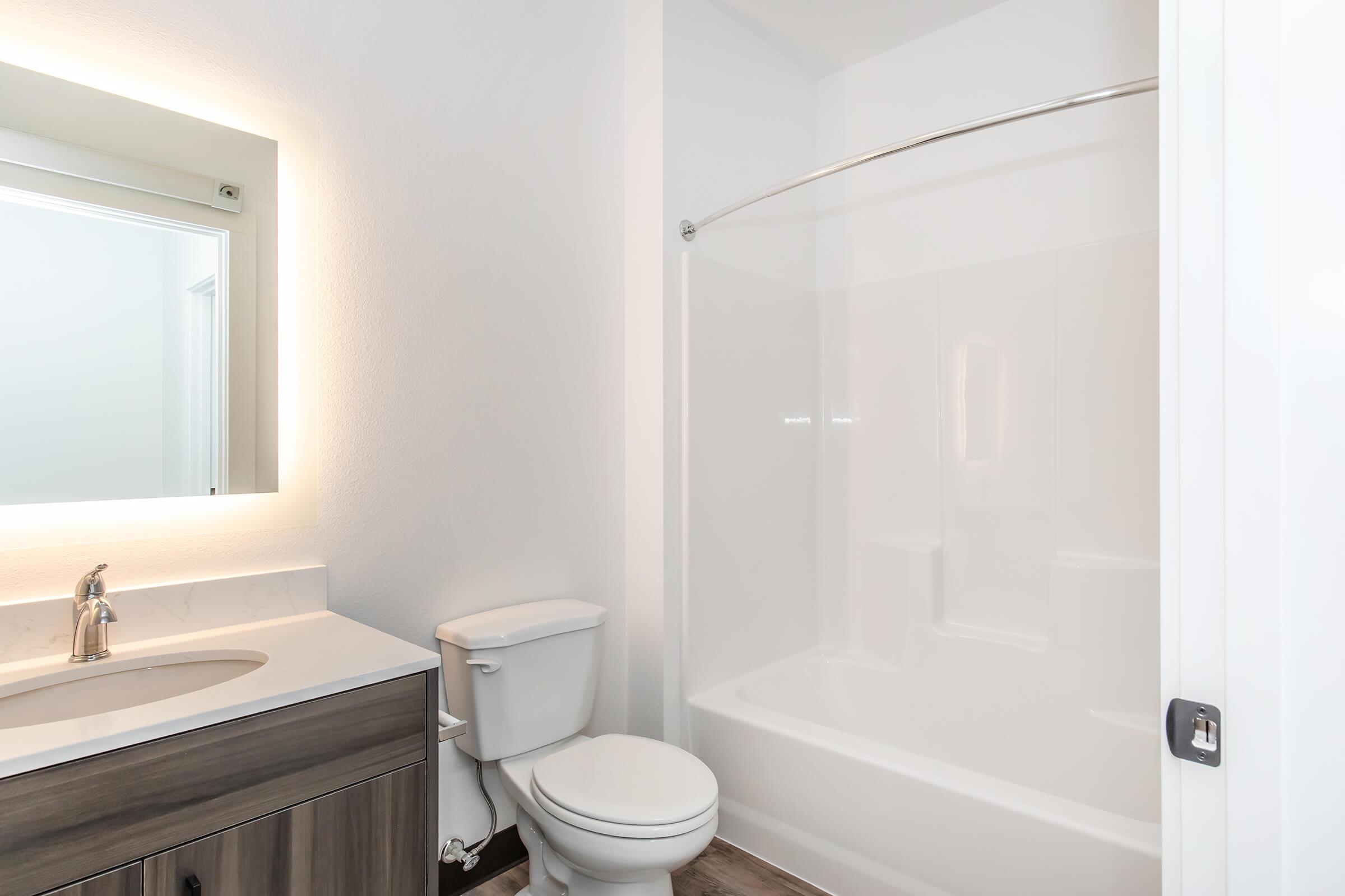 A modern bathroom featuring a clean design with a white bathtub and shower combo, a toilet, and a stylish vanity with a sink. The space is well-lit with a backlit mirror and has light-colored walls and a wooden floor, creating a bright and inviting atmosphere.