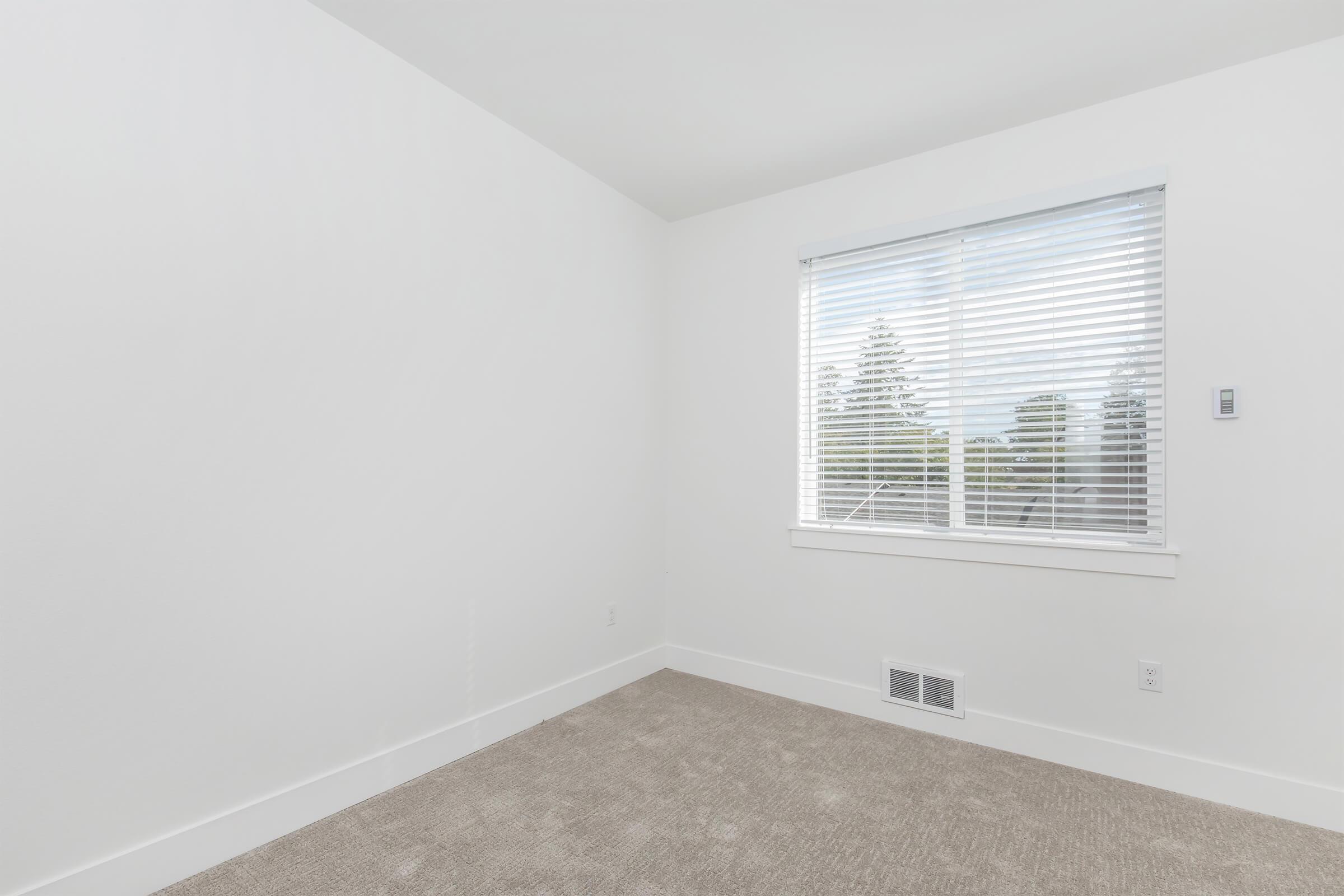 Empty room with light-colored walls and a large window featuring white blinds. The floor is covered in beige carpet. There is a vent on the wall and natural light is coming through the window, illuminating the space.