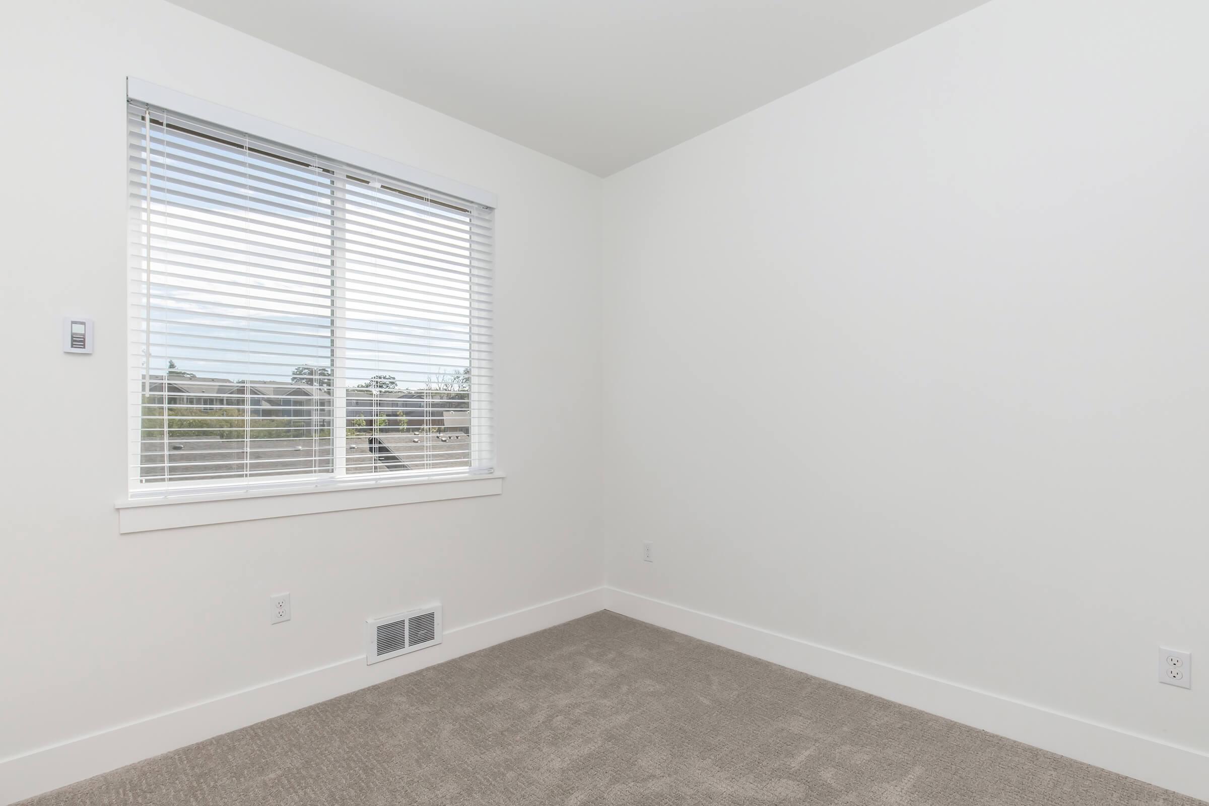 A small, empty room featuring white walls and light beige carpet. There is a window with white blinds on one wall, allowing natural light to enter. The opposite wall has a small vent and a light switch. The overall atmosphere is bright and minimalistic.