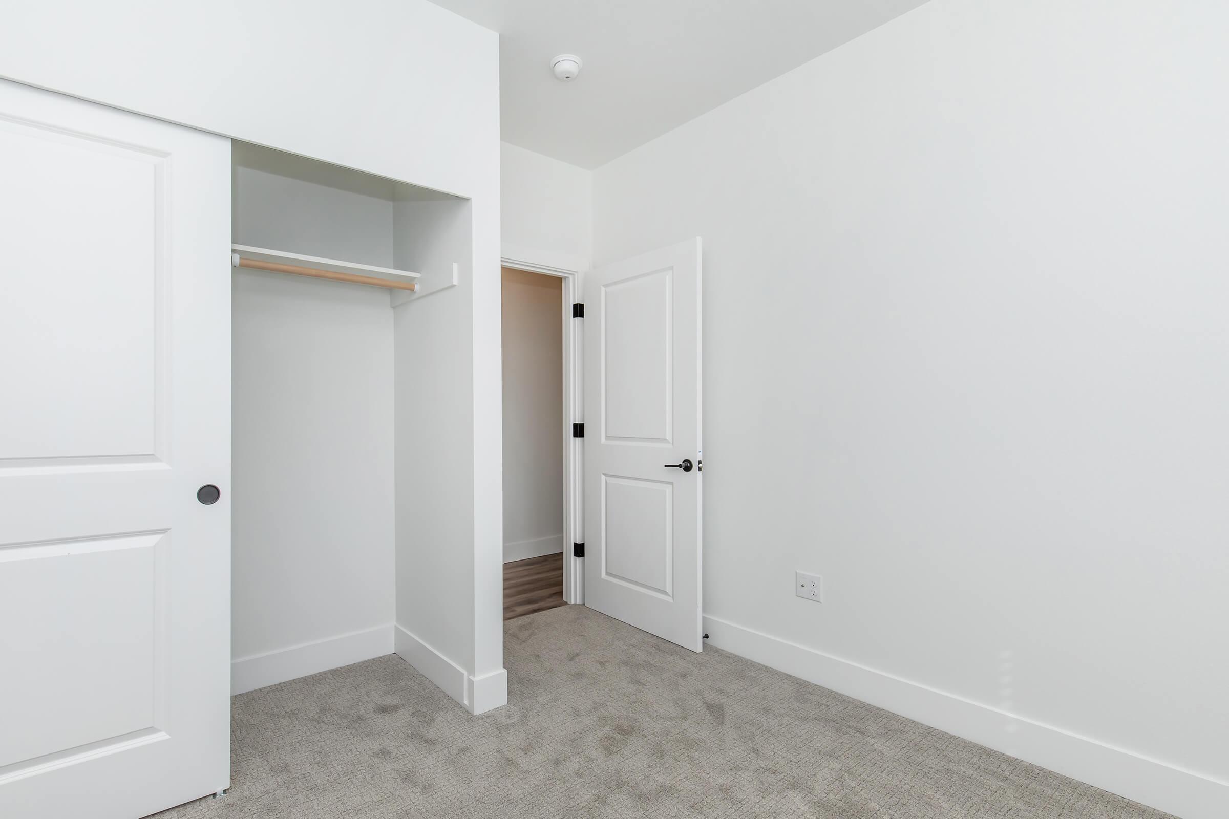 A minimalistic, empty room with light gray carpet, featuring a white closet with a wooden shelf, a closed door leading to another room, and a white wall. The room has a clean and modern design with natural light illuminating the space.