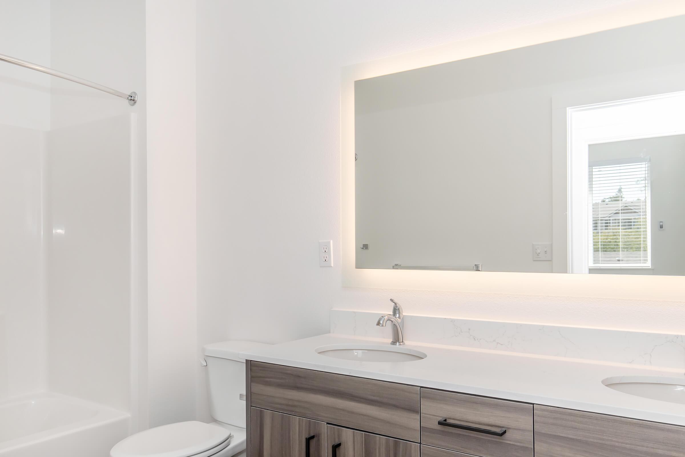 A modern bathroom featuring a double sink vanity with a sleek white countertop, a large mirror with soft lighting, and a separate shower area. The design includes light-colored walls and wooden cabinetry, creating a clean and contemporary aesthetic.