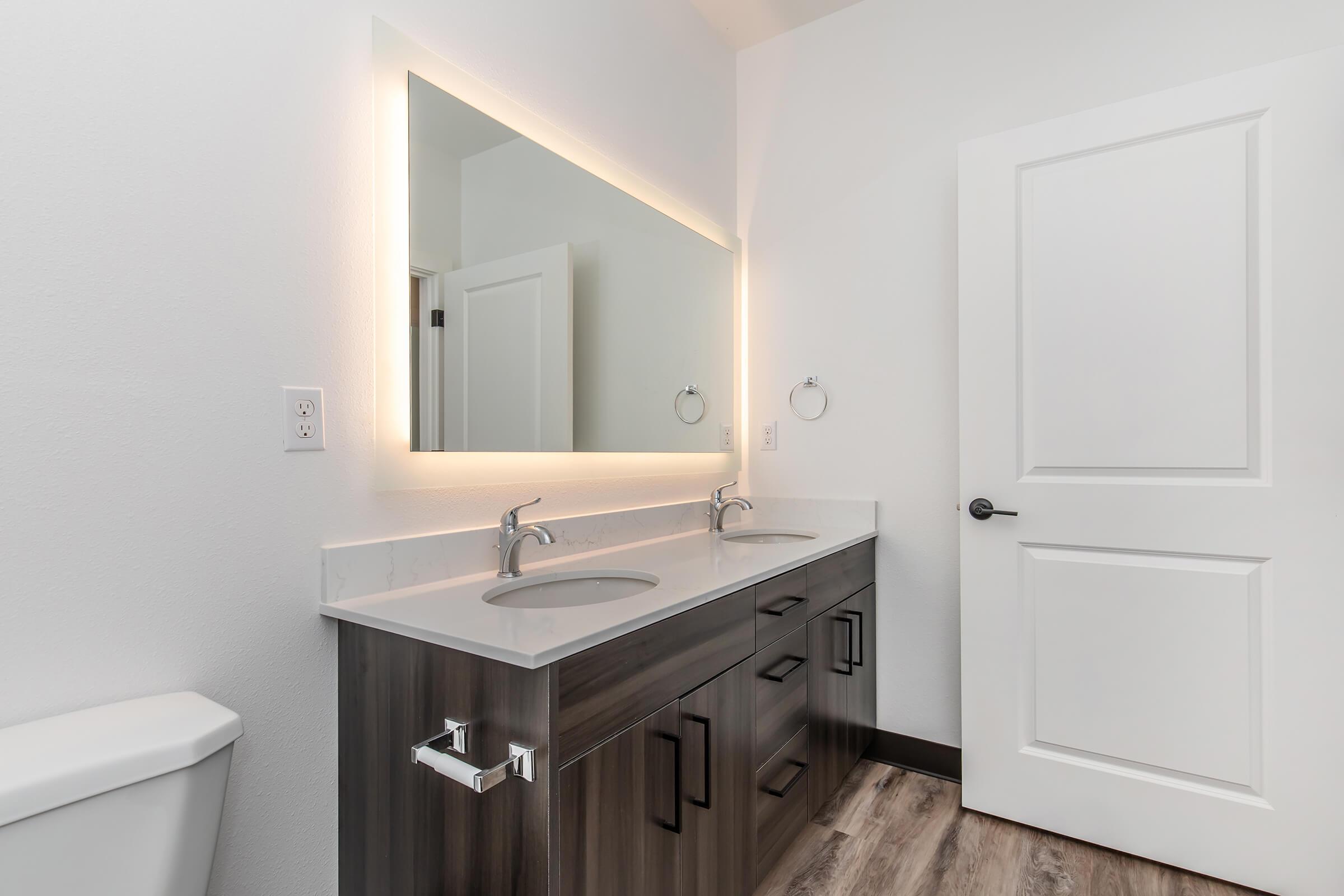 A modern bathroom featuring a dual sink vanity with brushed metal faucets, a large illuminated mirror above the sink, and a white door leading to another room. The walls are painted white, and the floor has a wood-like appearance. A toilet is visible on the left side of the image.