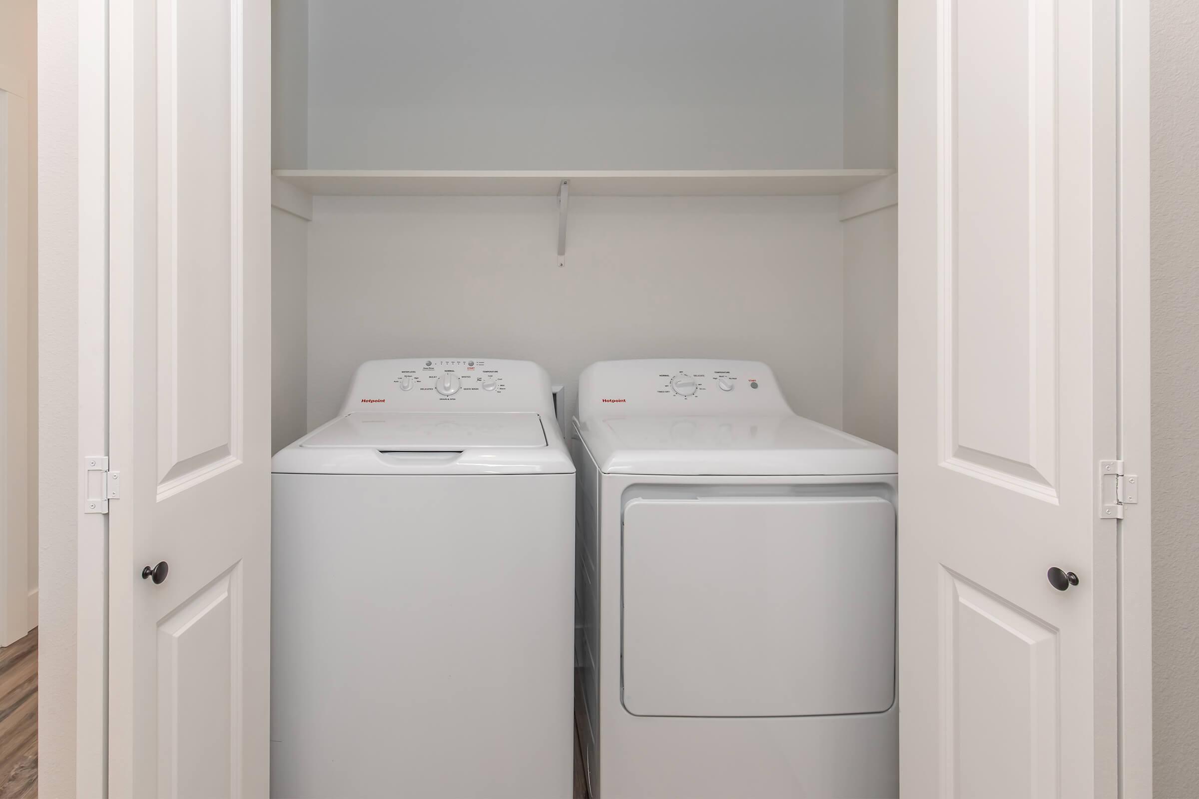 A compact laundry area featuring a stacked washer and dryer set, neatly enclosed within white cabinet doors. The space is well-lit with a neutral-colored wall and a shelf above the appliances for added storage. The flooring is a light wood finish, contributing to a clean and organized look.