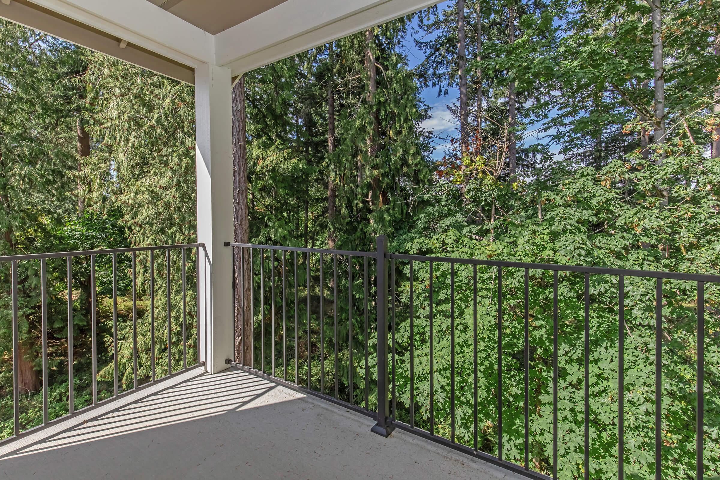 A view from a balcony overlooking a dense green forest. The balcony features black metal railings, and the surrounding trees create a tranquil, natural backdrop. Sunlight filters through the leaves, suggesting a bright day.