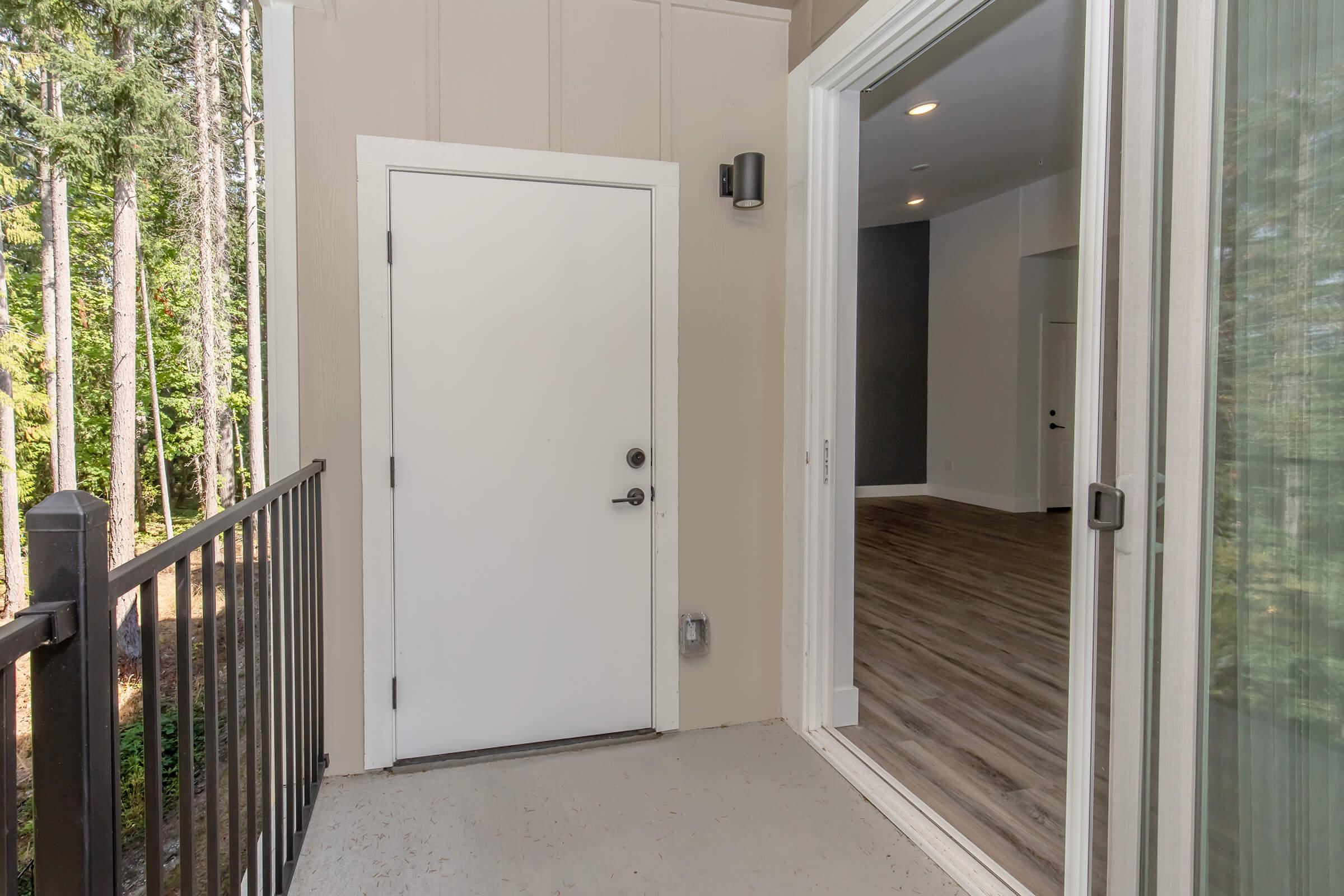 A doorway leading to a room with large windows, showcasing a view of tall trees outside. The space features a modern interior with light-colored walls and wood flooring. The door is white, and there is a balcony railing visible in the foreground. Natural light streams into the room.