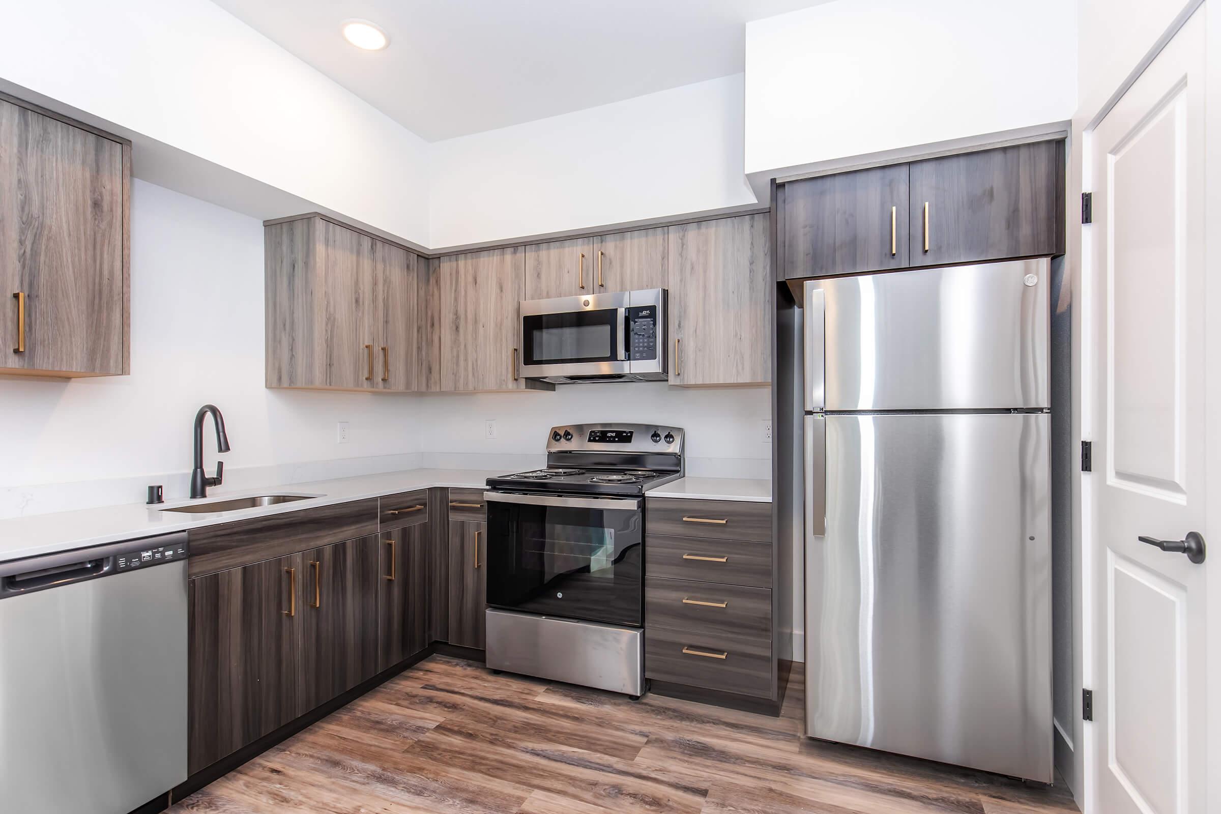 Modern kitchen featuring a mix of light and dark wooden cabinets, stainless steel appliances including a microwave, stove, and refrigerator. The countertop is sleek, and a single-basin sink is present, with ample storage space and a contemporary design aesthetic. Natural light enhances the open layout.