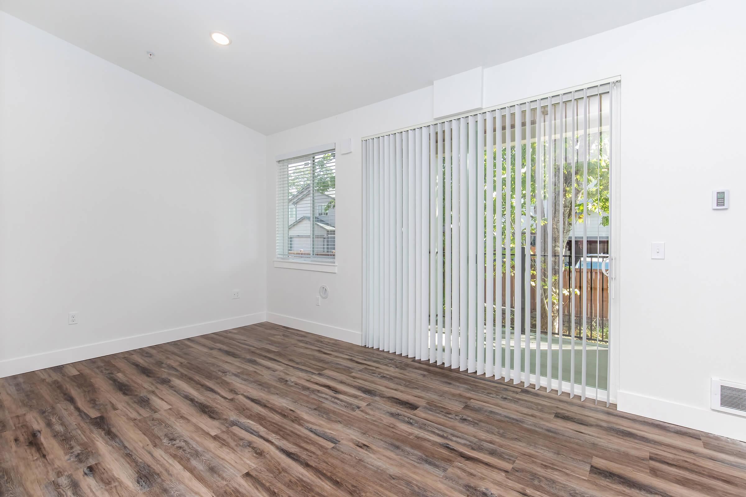 A bright, empty living room featuring wood-style flooring, white walls, and vertical blinds covering a large window leading to an outdoor area. Natural light is coming through the blinds, creating a warm atmosphere. The space is ready for furnishings and personal decoration.