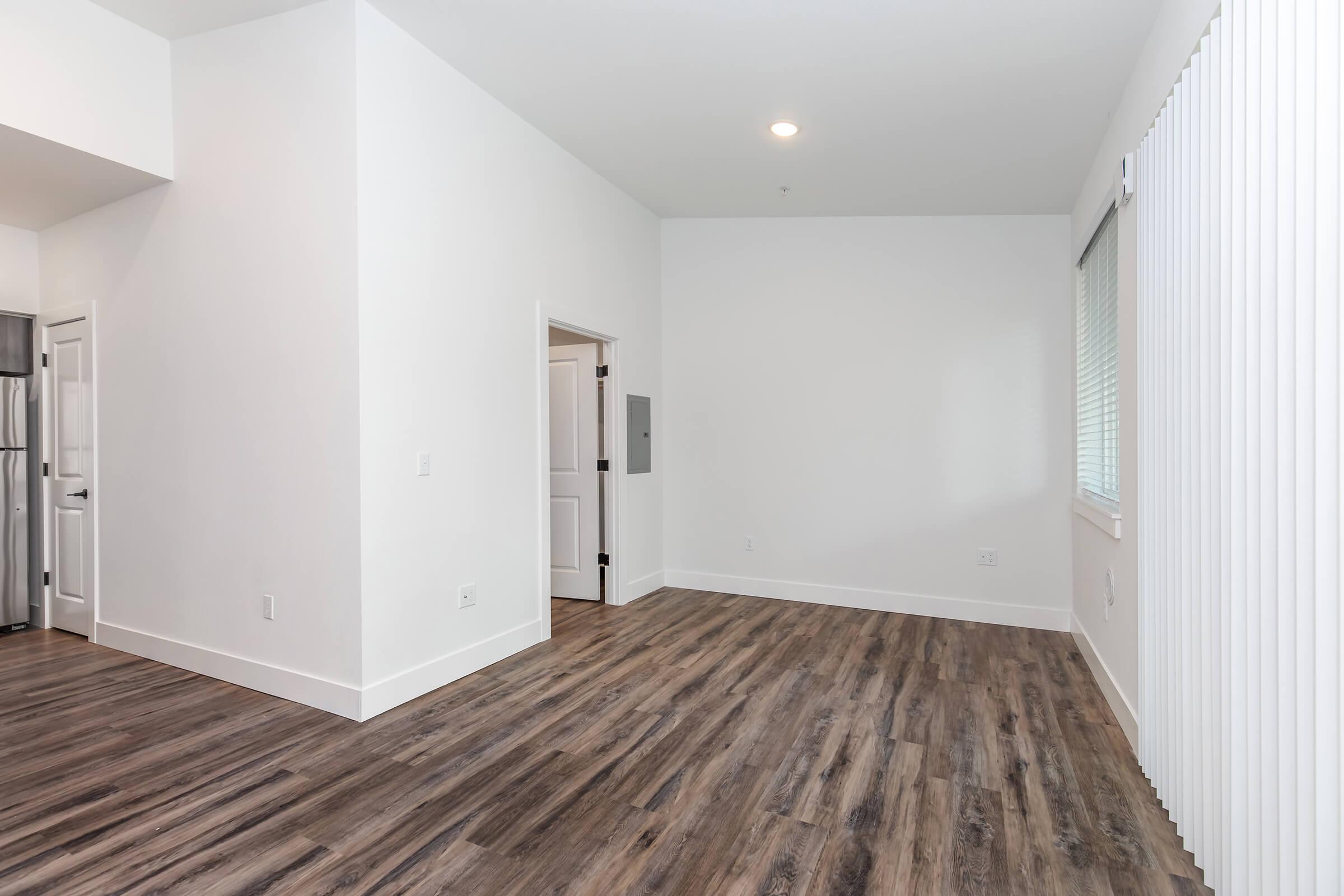 A spacious, empty room with white walls and a large window featuring vertical blinds. The floor is made of brown wood-like tiles. A door on the left leads to another area, and there is a slight reflection of light from a ceiling fixture. The overall atmosphere is bright and minimalistic.