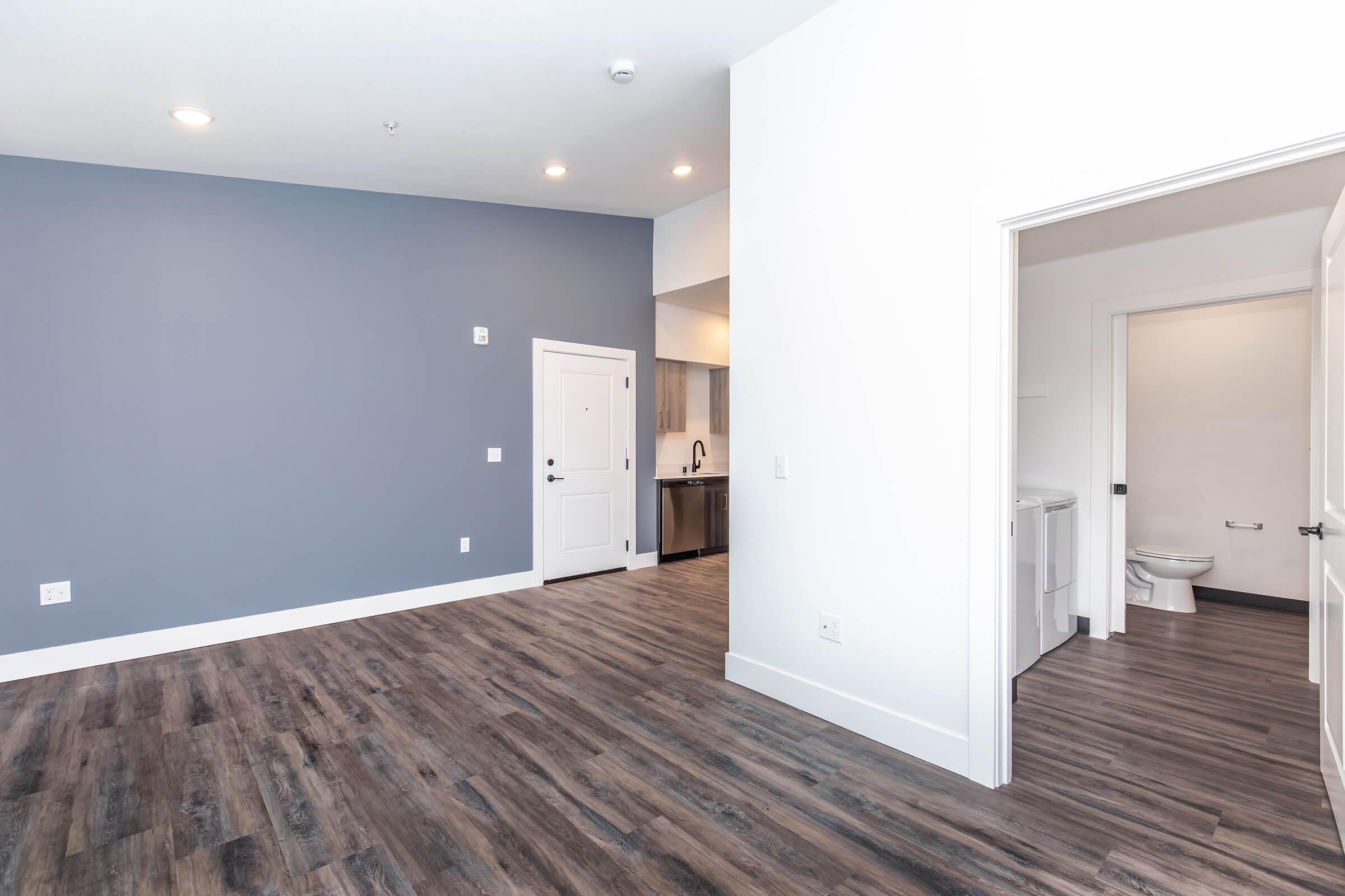 A spacious, modern room featuring light-colored walls and a dark wood-style floor. The room includes a doorway leading to a kitchen area, with a glimpse of a bathroom visible in the background. The overall design is bright and minimalist, highlighting a clean aesthetic.