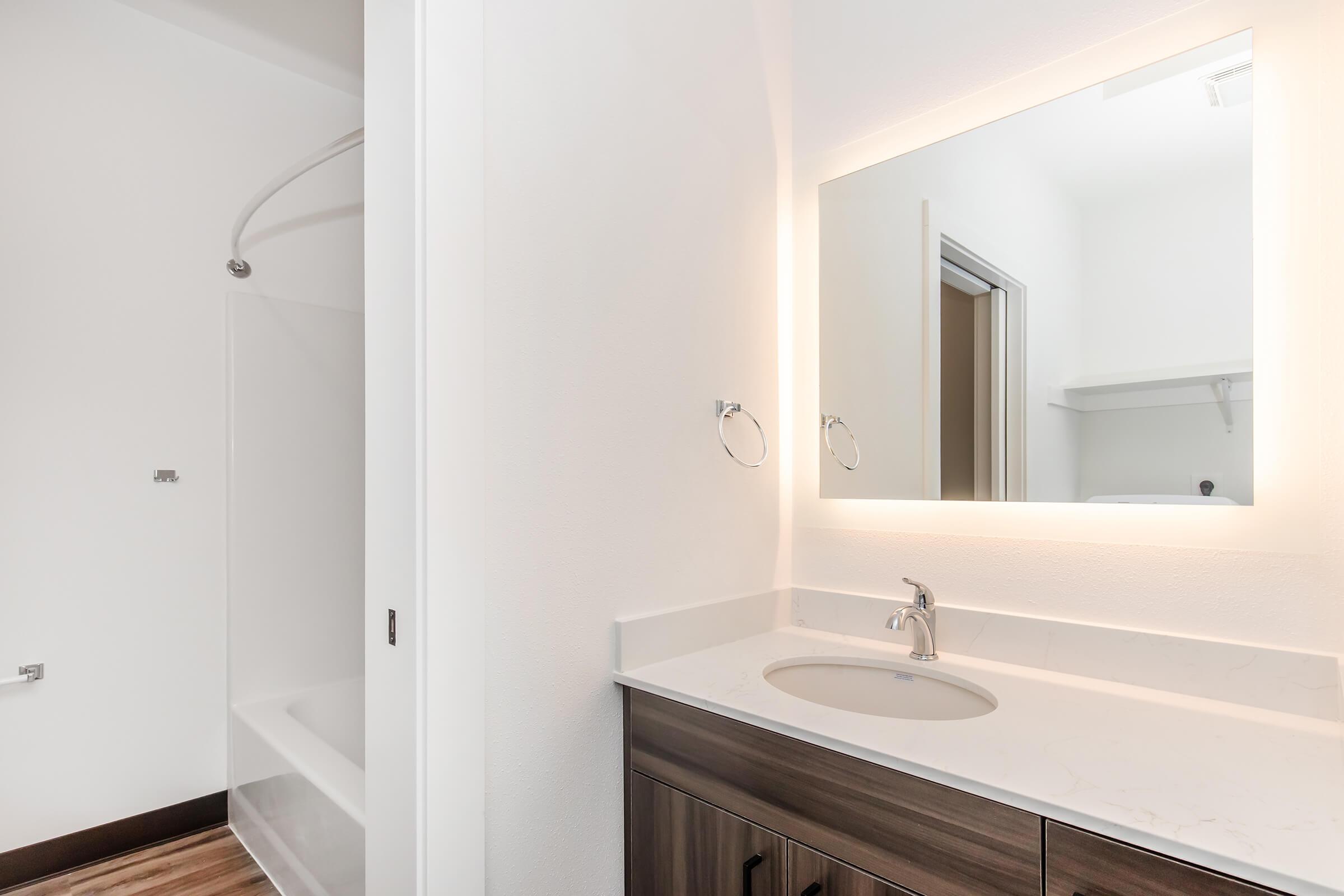 A modern bathroom featuring a shower and bathtub combo, a sleek vanity with a white countertop, and a large illuminated mirror. The walls are painted white and the floor has a wood-like design, creating a clean and contemporary atmosphere.