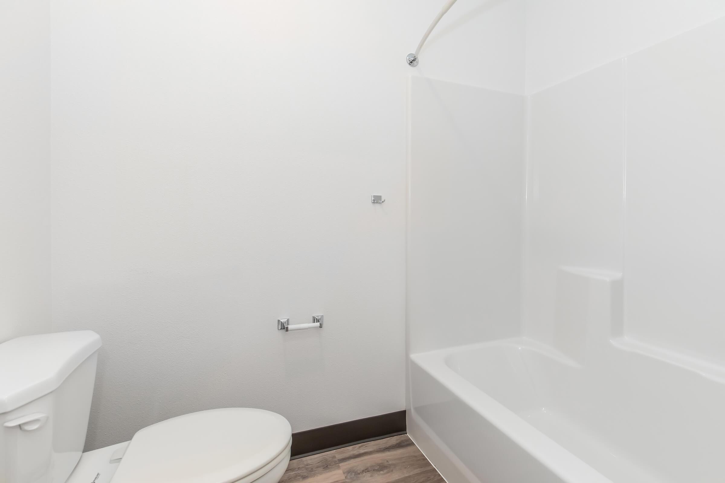 A clean bathroom featuring a white toilet and a bathtub with a shower. The walls are painted white, and the floor is a light wooden surface. The bathroom has minimal fixtures, including a toilet paper holder near the toilet. Overall, it presents a simple and modern aesthetic.
