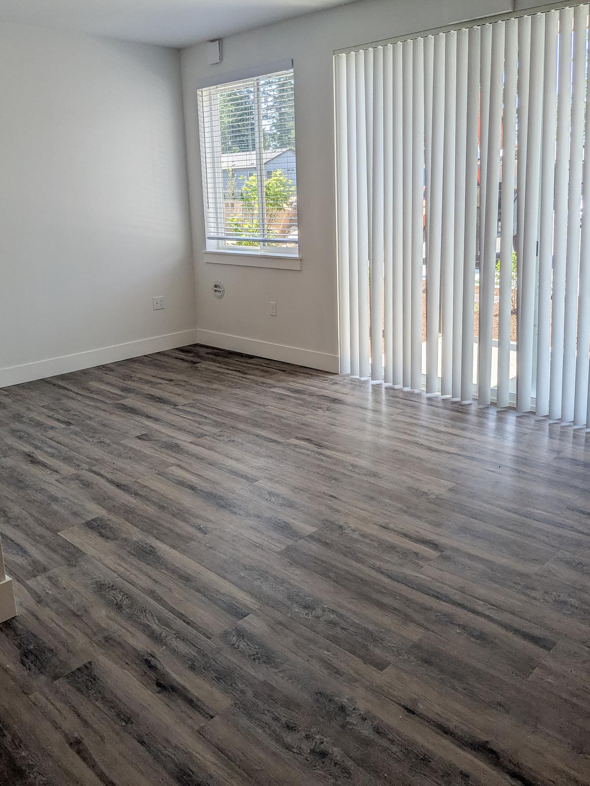 A spacious, empty room with light-colored walls and dark wood-like flooring. There is a large window with vertical blinds allowing natural light to enter, and a sliding glass door to the outside. The room appears clean and modern, perfect for furnishing and personalizing.