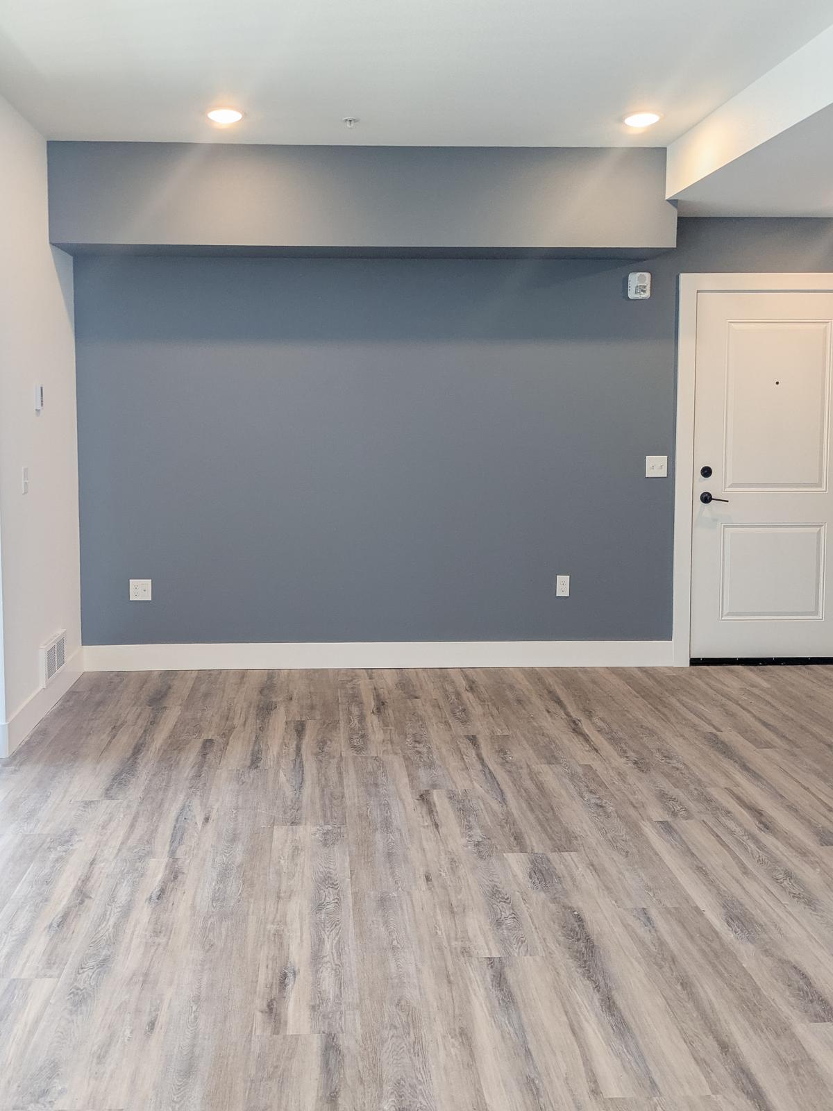 Empty room with light-colored laminate flooring and a gray accent wall. A white door with a knob is visible on the right side, and the room is well-lit with recessed lighting in the ceiling. There are no furnishings or decorations in the space.