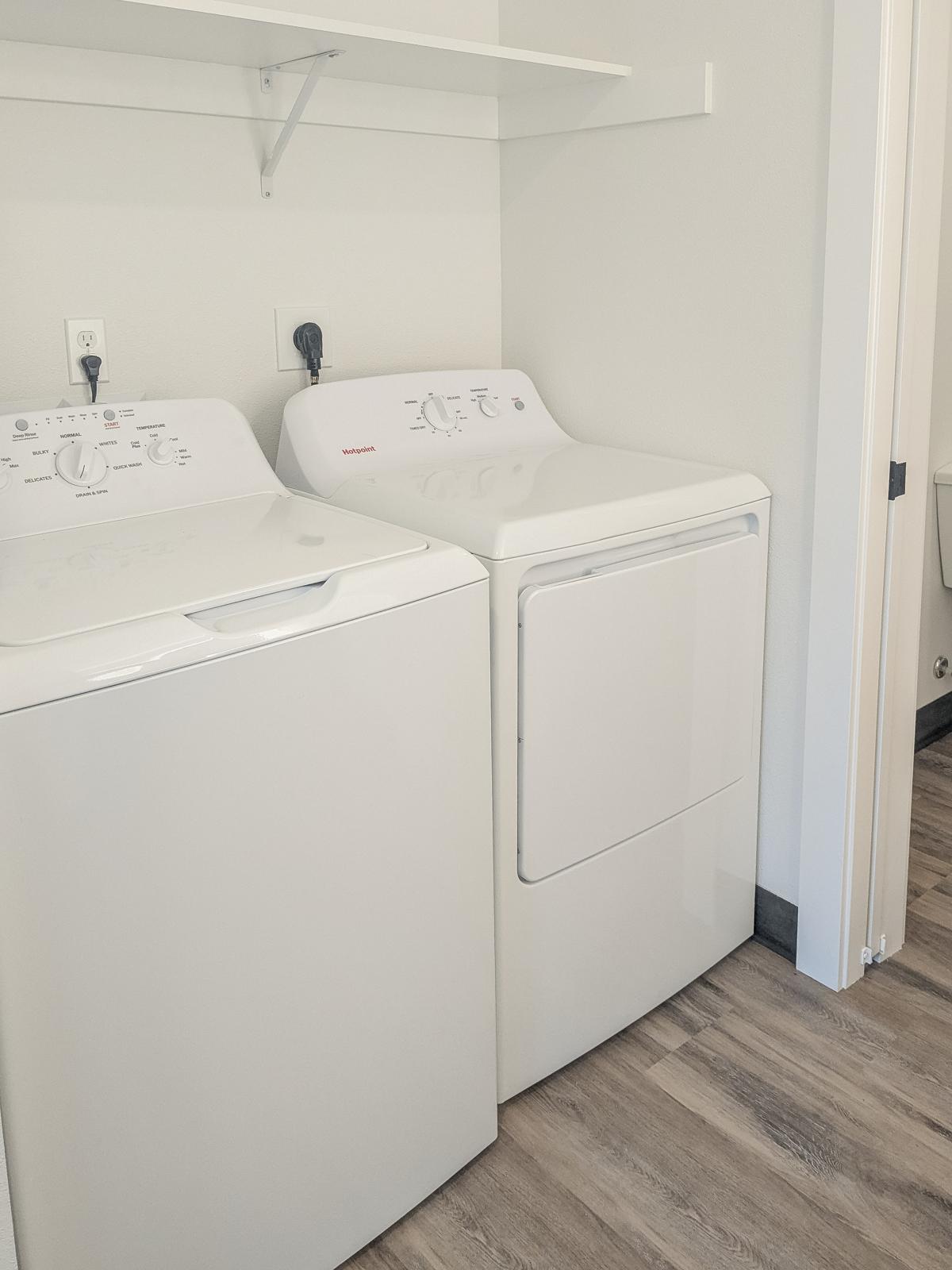 A clean laundry room featuring a white washing machine and a white dryer side by side on a wooden floor. The wall is painted light gray, and there's a shelf above the appliances. A doorway is visible in the background, indicating access to another room.