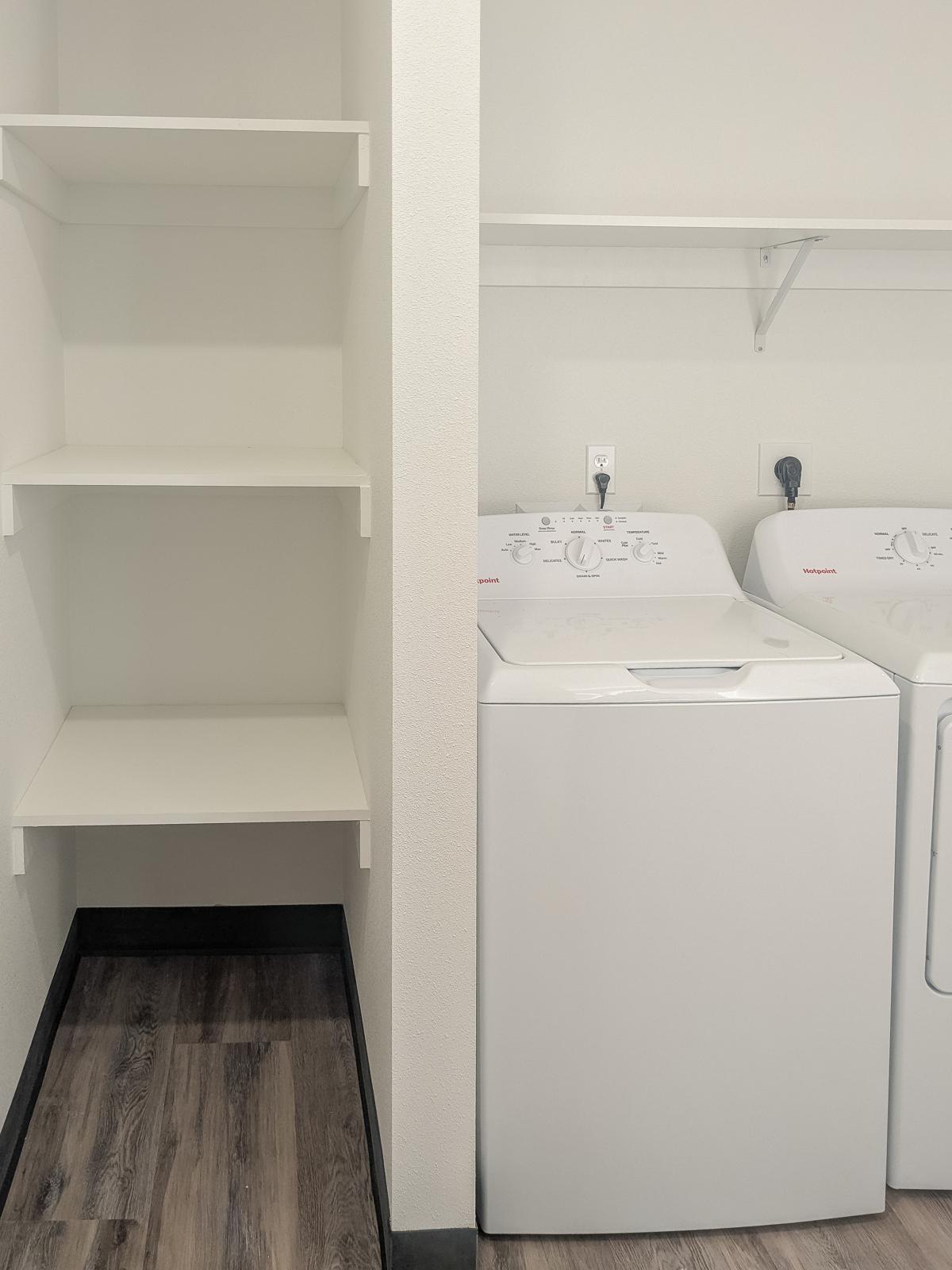 A laundry area featuring a white washing machine and dryer side by side, with a clean, minimalist design. To the left, there are empty white shelves mounted on the wall, and the flooring is a dark laminate. The walls are painted a light color, creating a bright and airy atmosphere.