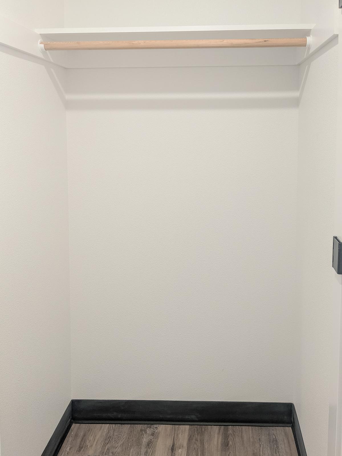 Empty closet space with a single wooden rod for hanging clothes. The walls are plain white, and the floor features dark, wooden-looking planks. There is no clutter or items present in the closet.