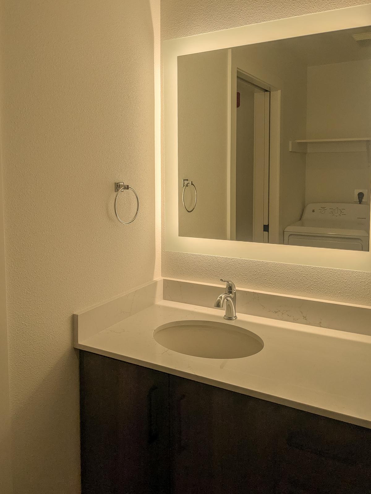 A modern bathroom featuring a lighted mirror above a sink with a marble countertop. The walls are painted in a light color, and there is a towel ring on the wall. A door to another room is partially visible, along with a stacked washer and dryer in the background. The overall atmosphere is clean and minimalistic.