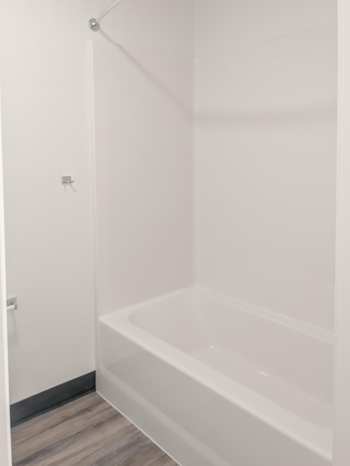 A clean, modern bathroom featuring a white bathtub with a shower head above. The walls are smooth and light-colored, and the floor is covered with wooden-style laminate flooring. There is minimal decor, creating a simple and uncluttered look.