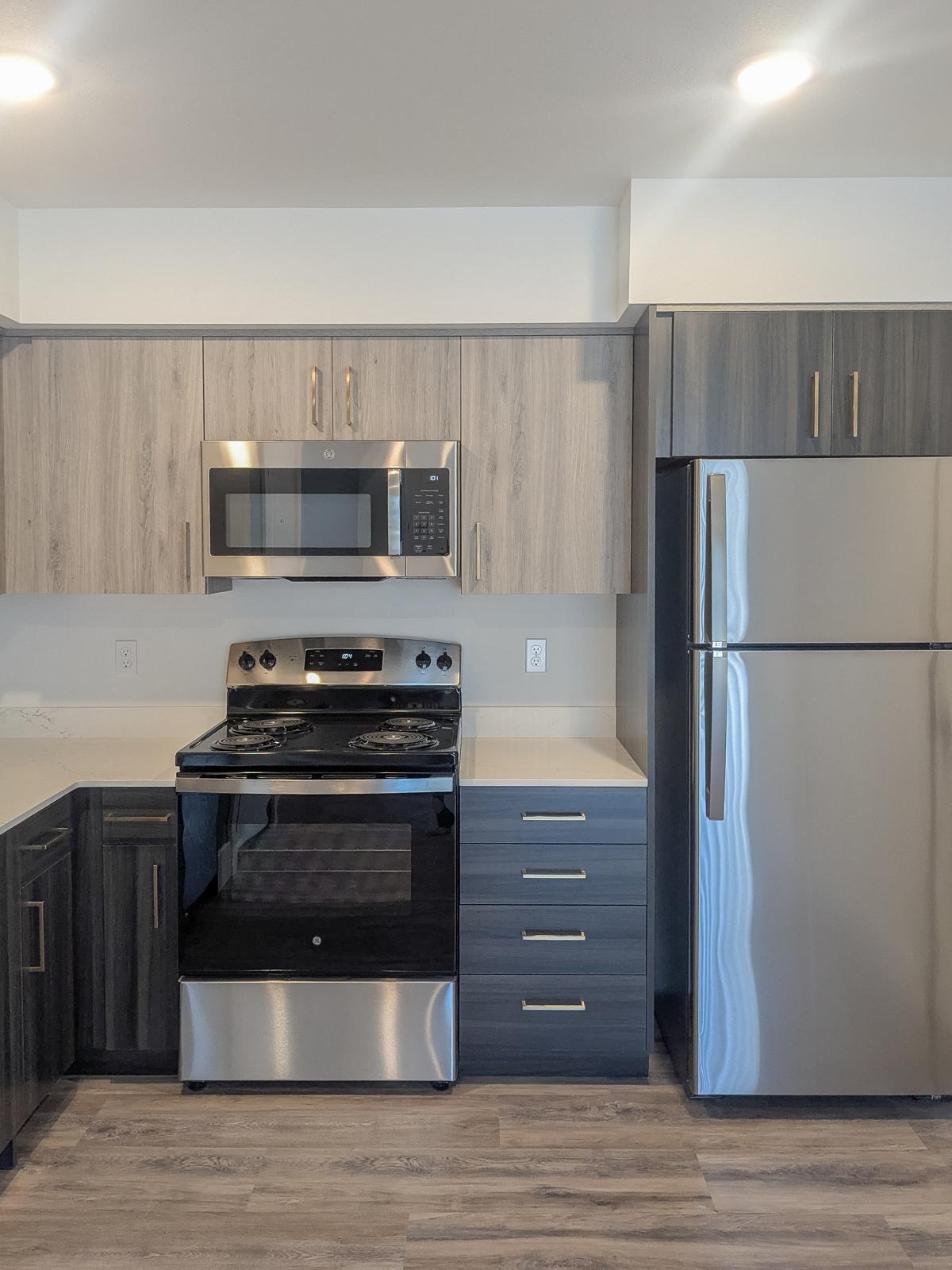 Modern kitchen featuring stainless steel appliances, including a microwave, stove, and refrigerator. The cabinetry has a wood grain finish with sleek, dark colors. The countertop is light-colored, and the flooring consists of laminate planks, creating a contemporary and stylish look.