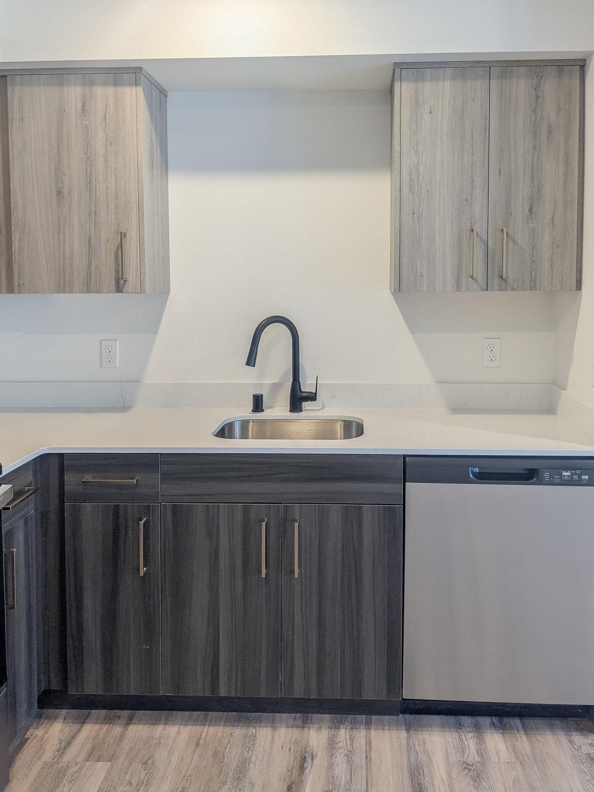 Modern kitchen with gray wood cabinetry, a stainless steel sink, and a dishwasher. The countertop is light-colored, and the cabinetry features sleek gold handles. The design is minimalist and contemporary, with a clean aesthetic and a light wall background.